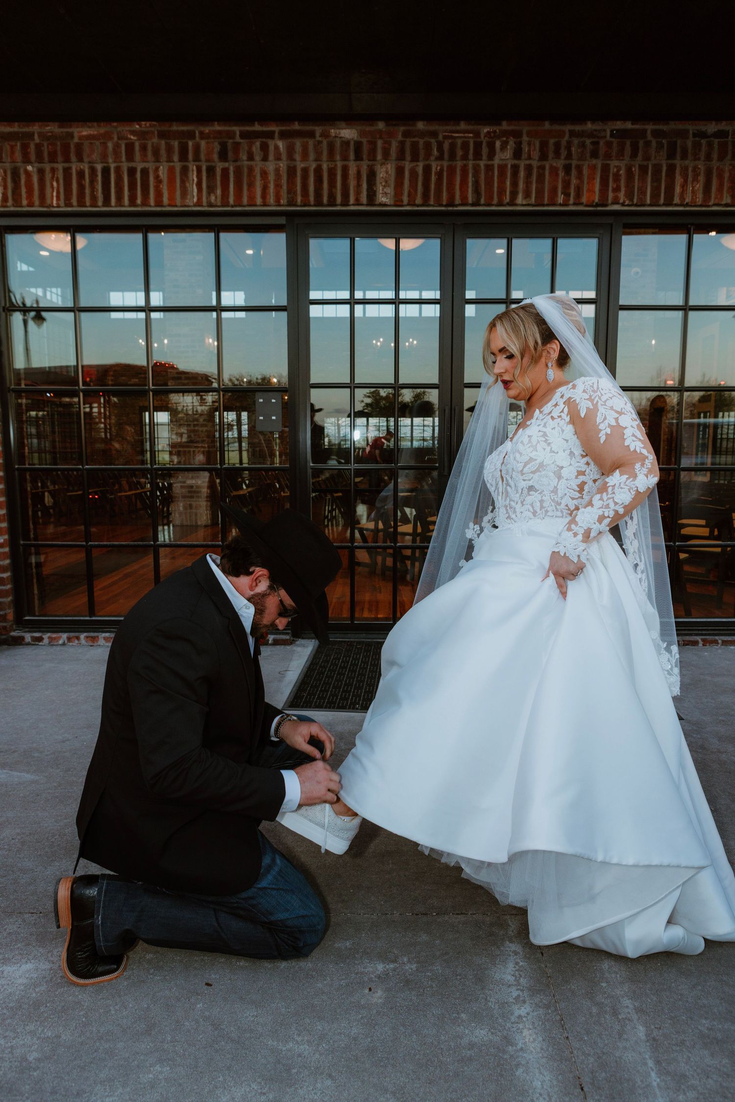 Groom kneeling to adjust bride's wedding dress shoe outdoors near industrial windows.
