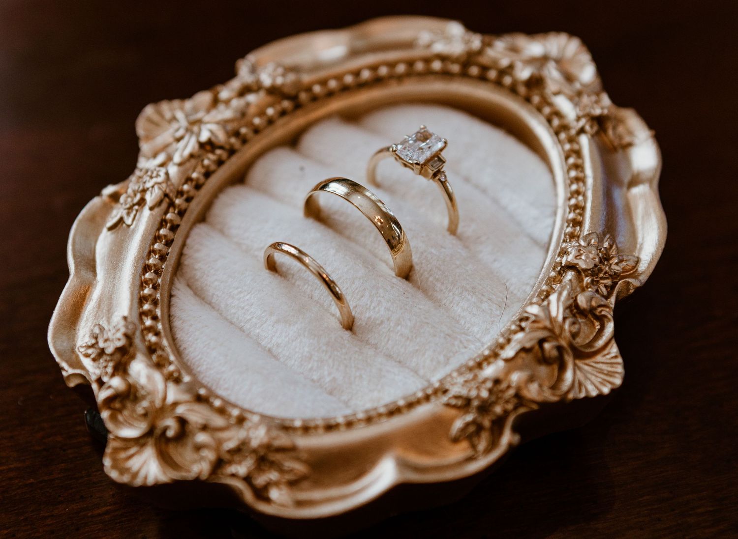 Three gold wedding rings displayed on a white velvet cushion in an ornate gold vintage frame on a dark wood surface.