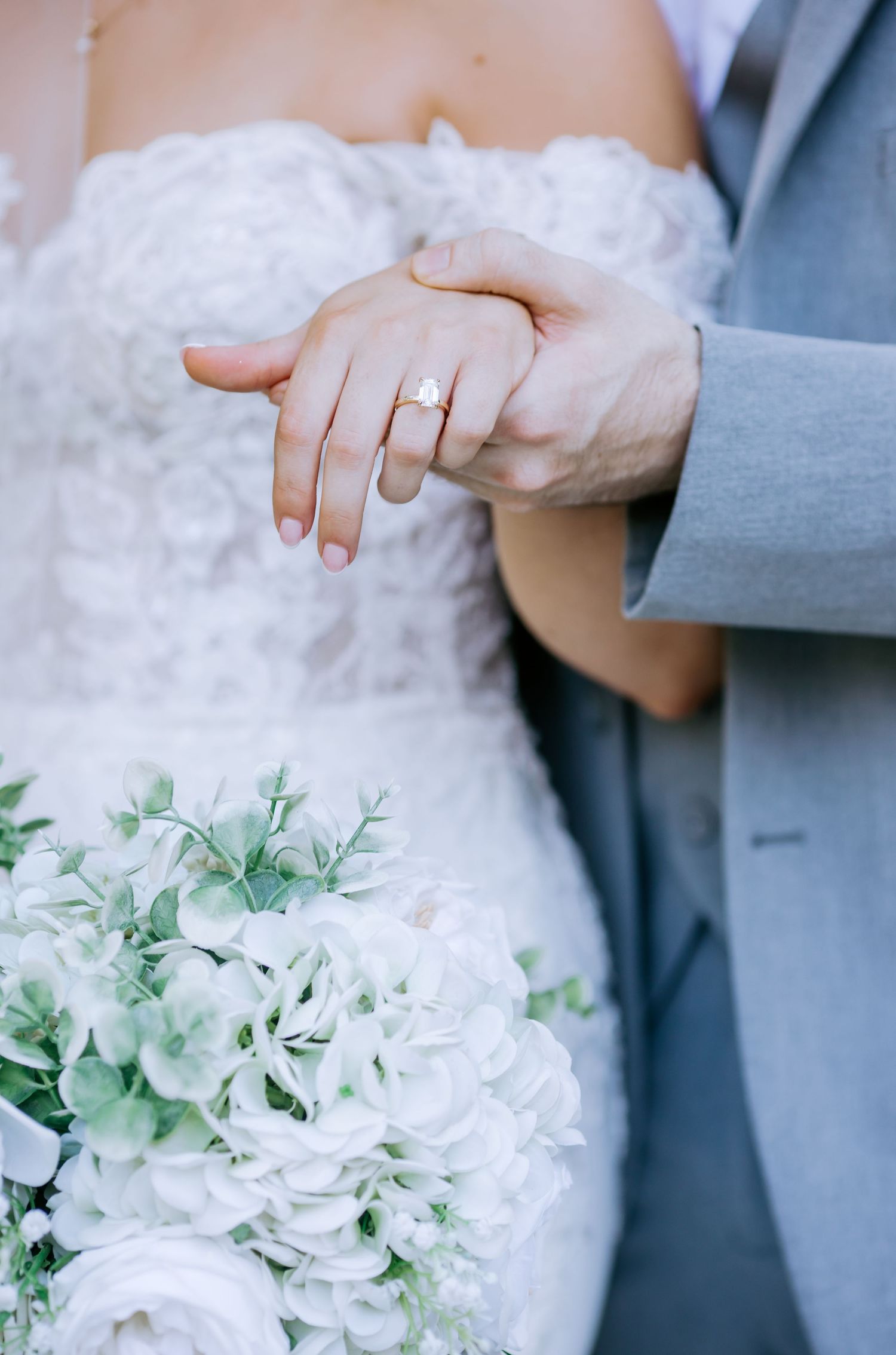 Bride and groom holding hands displaying diamond engagement ring with white floral bouquet at wedding ceremony.