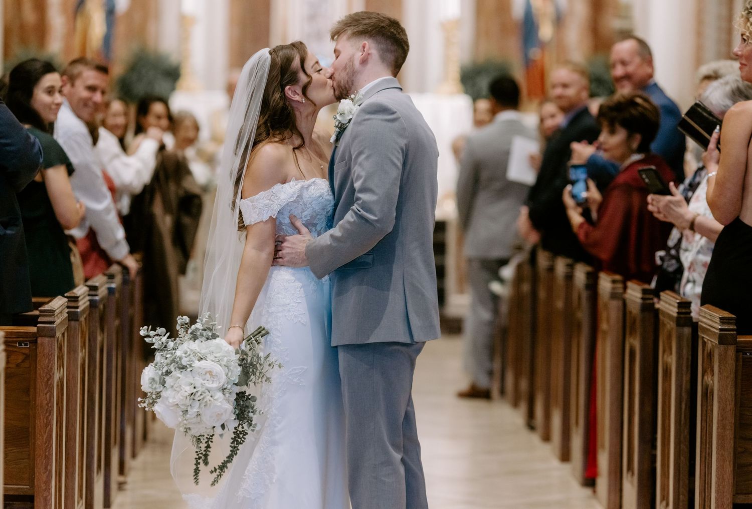 Bride and groom share first kiss walking down church aisle surrounded by cheering wedding guests.
