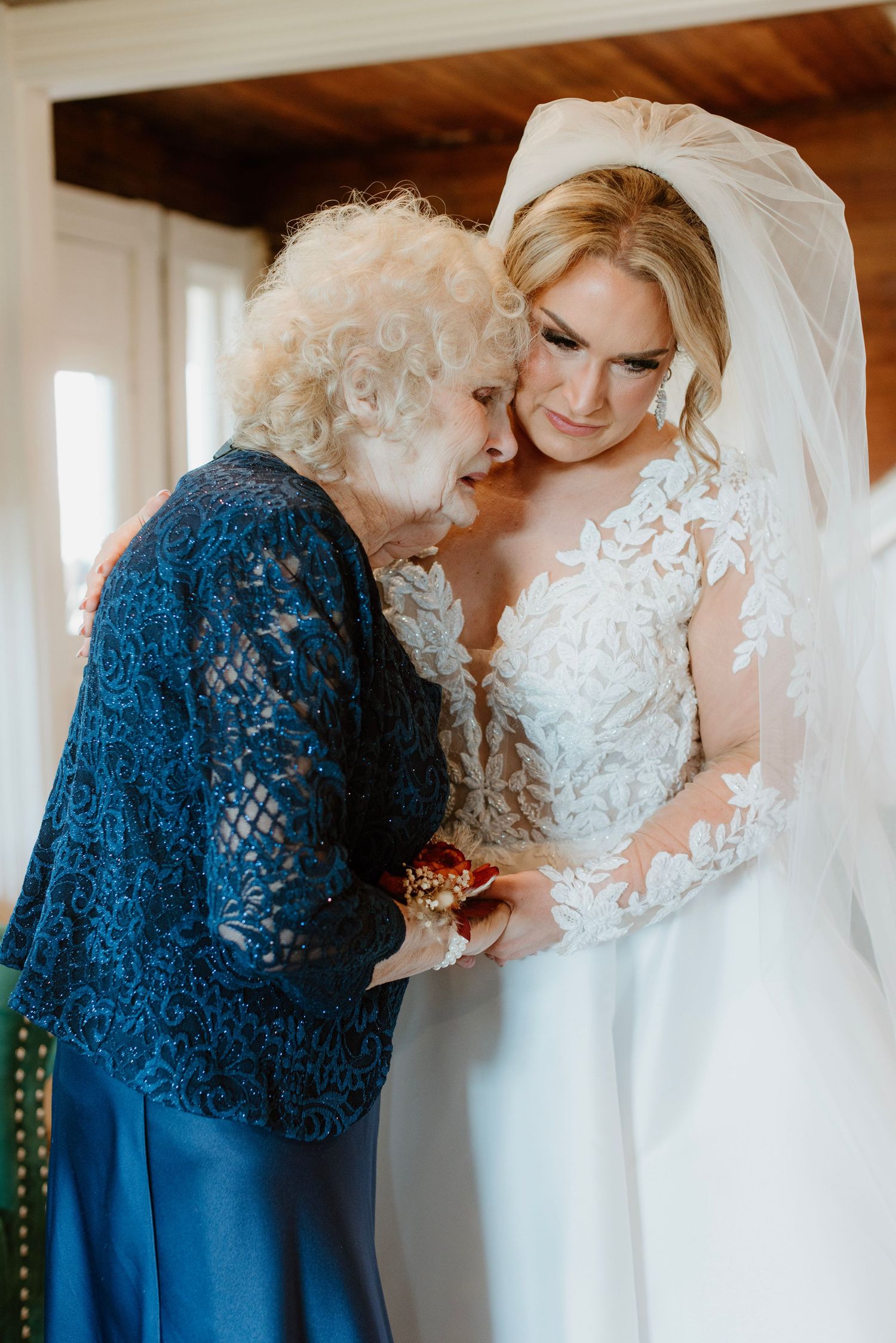 Grandmother in blue lace jacket helps bride in white gown with corsage.
