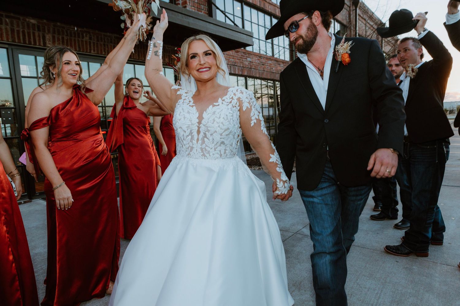 Bride in white lace gown walks with groom in cowboy hat as bridesmaids in red dresses celebrate.
