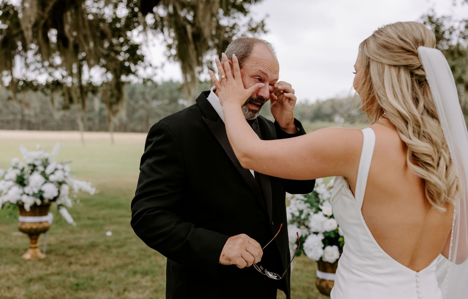 Emotional father in a dark suit wipes away tears while his daughter in a white wedding dress stands before him at an outdoor ceremony.