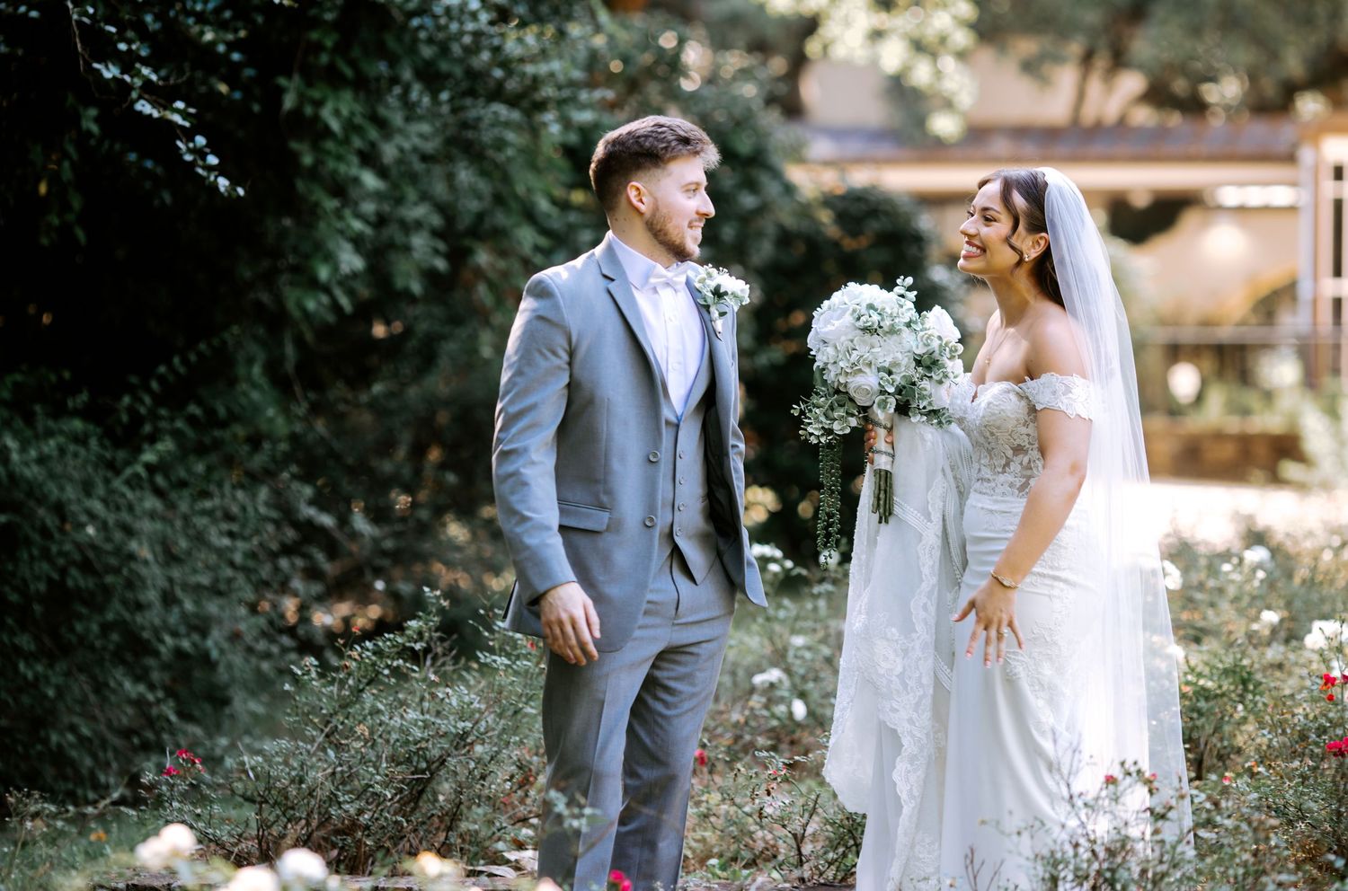Bride and groom stand facing each other in a lush garden, she holds a white bouquet in a lace gown with veil.