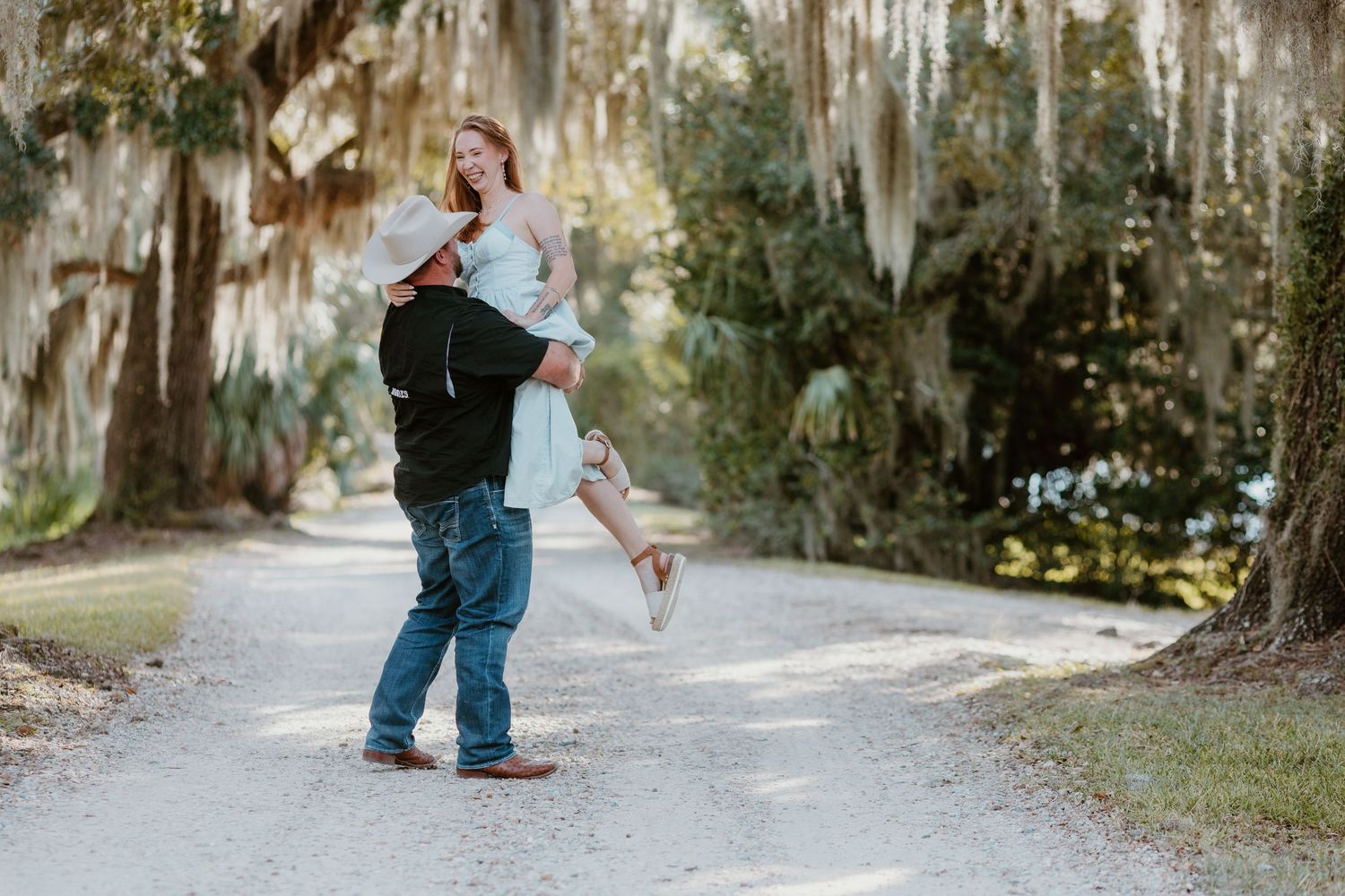 A couple dances playfully on a tree-lined path with Spanish moss hanging from branches overhead.