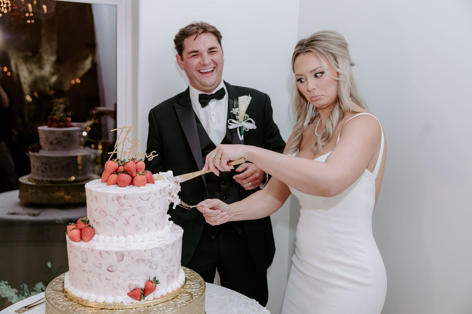 Smiling bride and groom cutting their white wedding cake topped with fresh strawberries during a reception celebration.