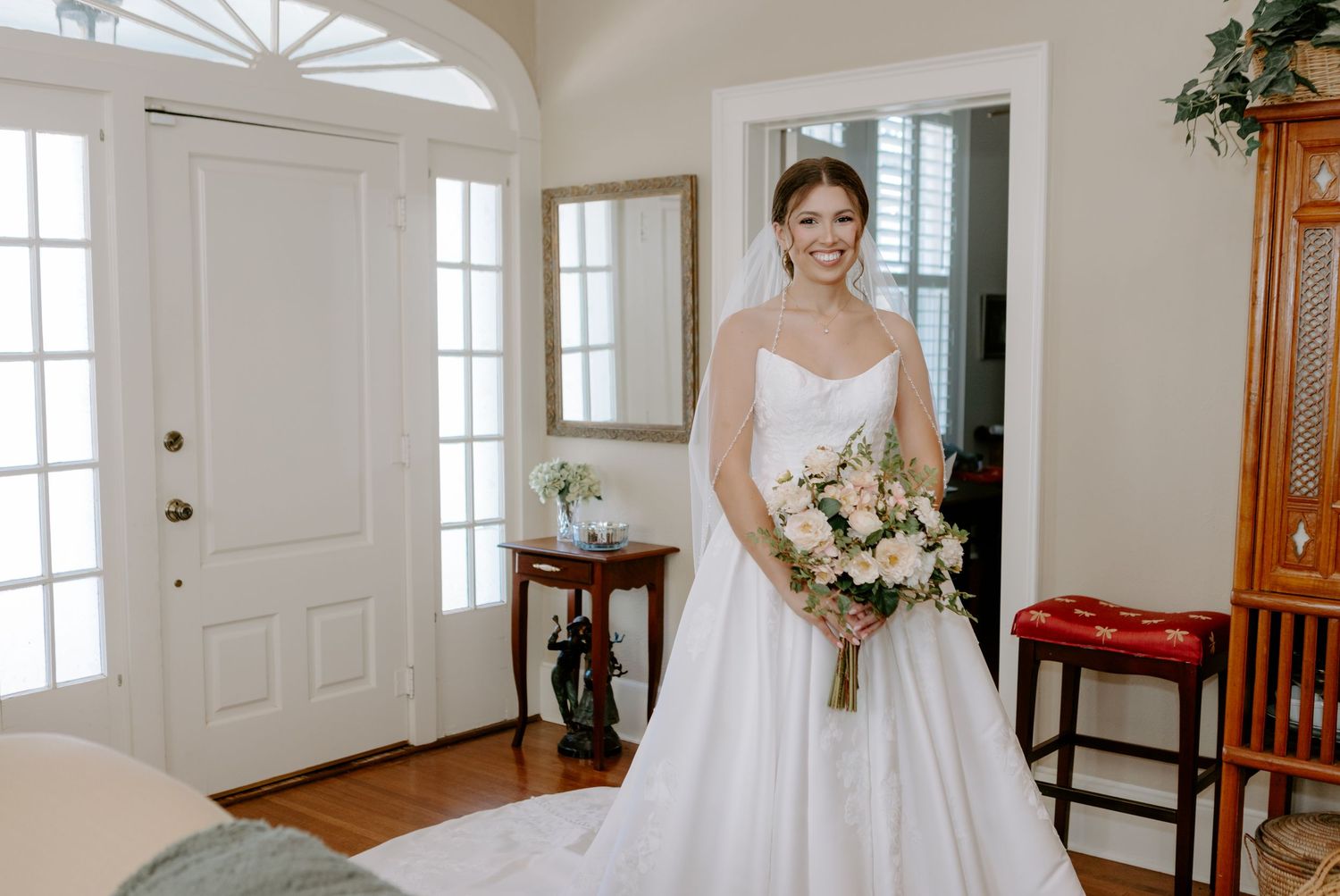 Smiling bride in a white strapless ball gown holding a pink floral bouquet standing in an elegant home entryway.