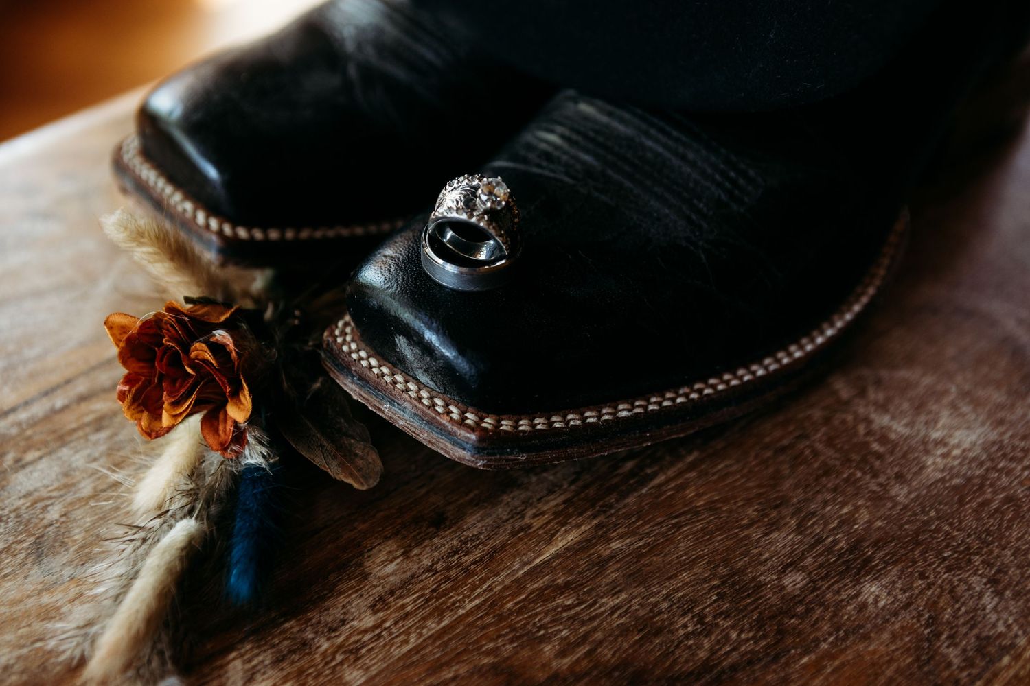 Black cowboy boots with silver rings and a rustic floral boutonniere on a wooden surface.