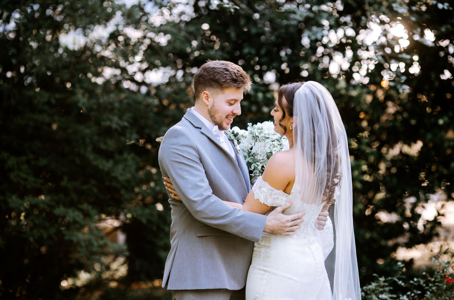 Groom in gray suit embracing bride in white lace gown and veil, sharing an intimate moment outdoors.