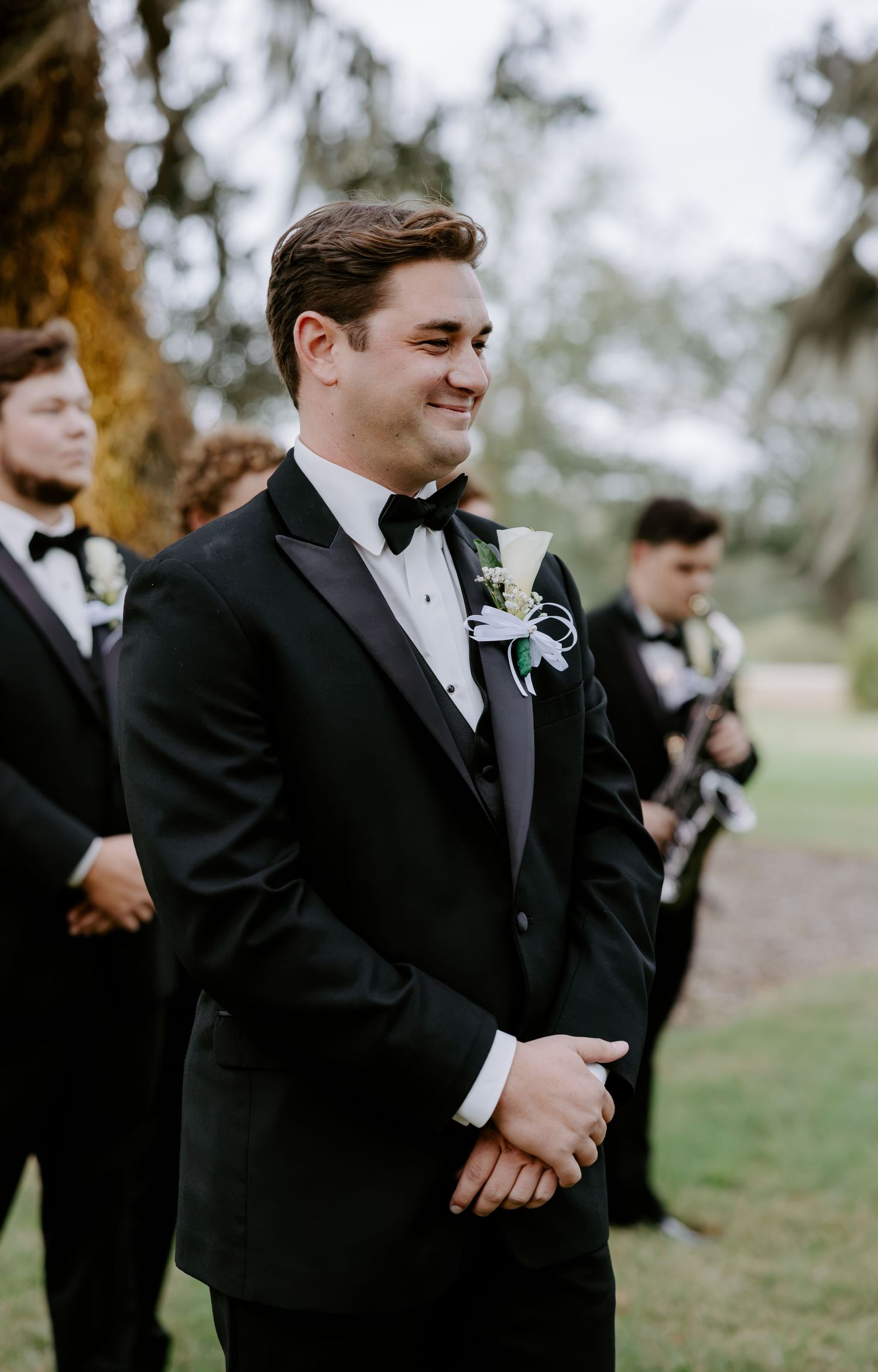 Smiling groom in a black tuxedo with a white boutonniere stands outdoors at a wedding ceremony with groomsmen in the background.