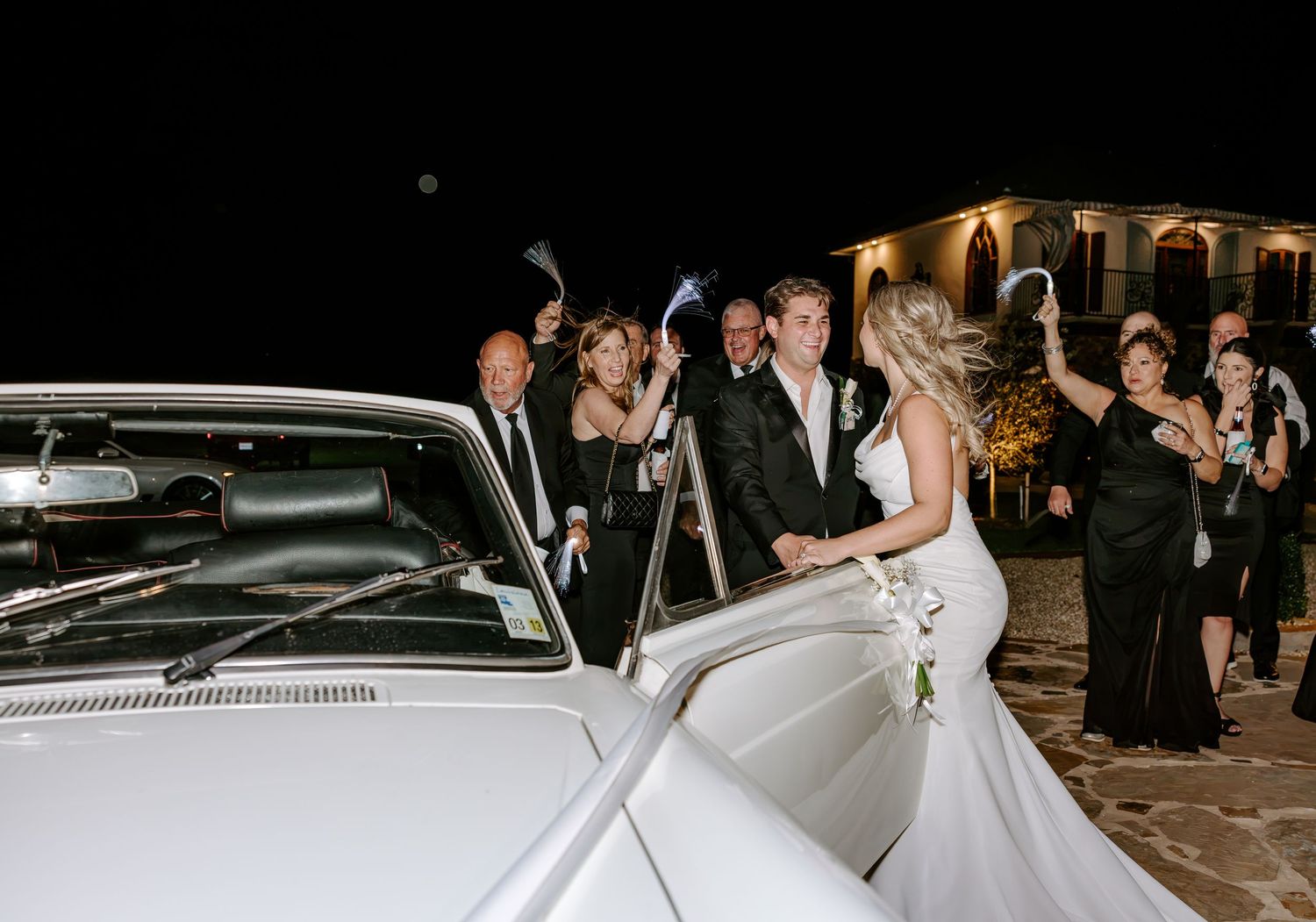 Bride and groom smile beside a vintage white car at night as wedding guests cheer and hold sparklers outside a lit venue.