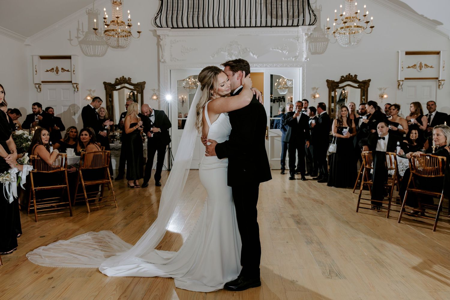 Bride in a long white gown and groom in a black suit share a first dance as wedding guests watch in an elegant chandelier-lit ballroom.