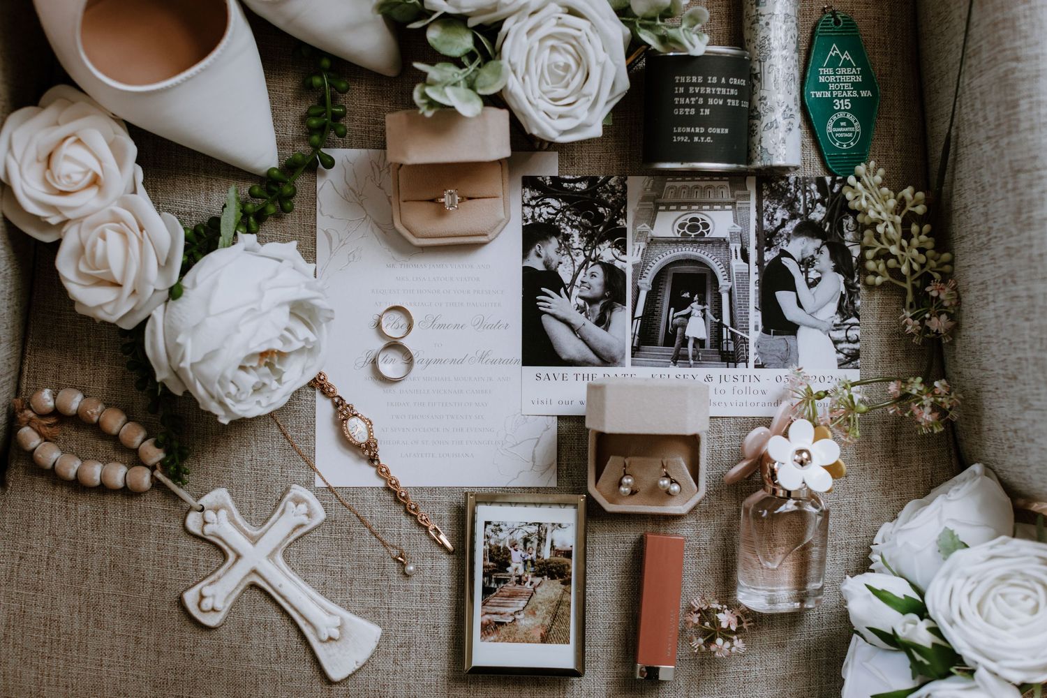 Elegant wedding flat lay with white roses, gold rings, cross, invitations, photos, perfume, and accessories on burlap.