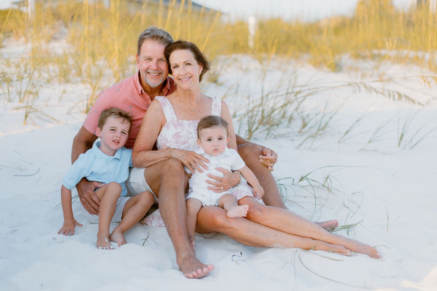 A family relaxes together on a white sandy beach during sunset with beach grass in the background.