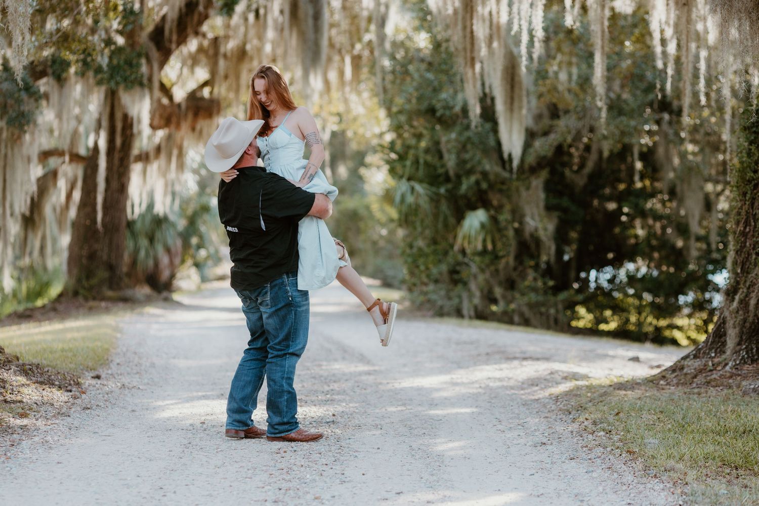 A person in denim overalls walks down a tree-lined path covered in Spanish moss on a sunny day.