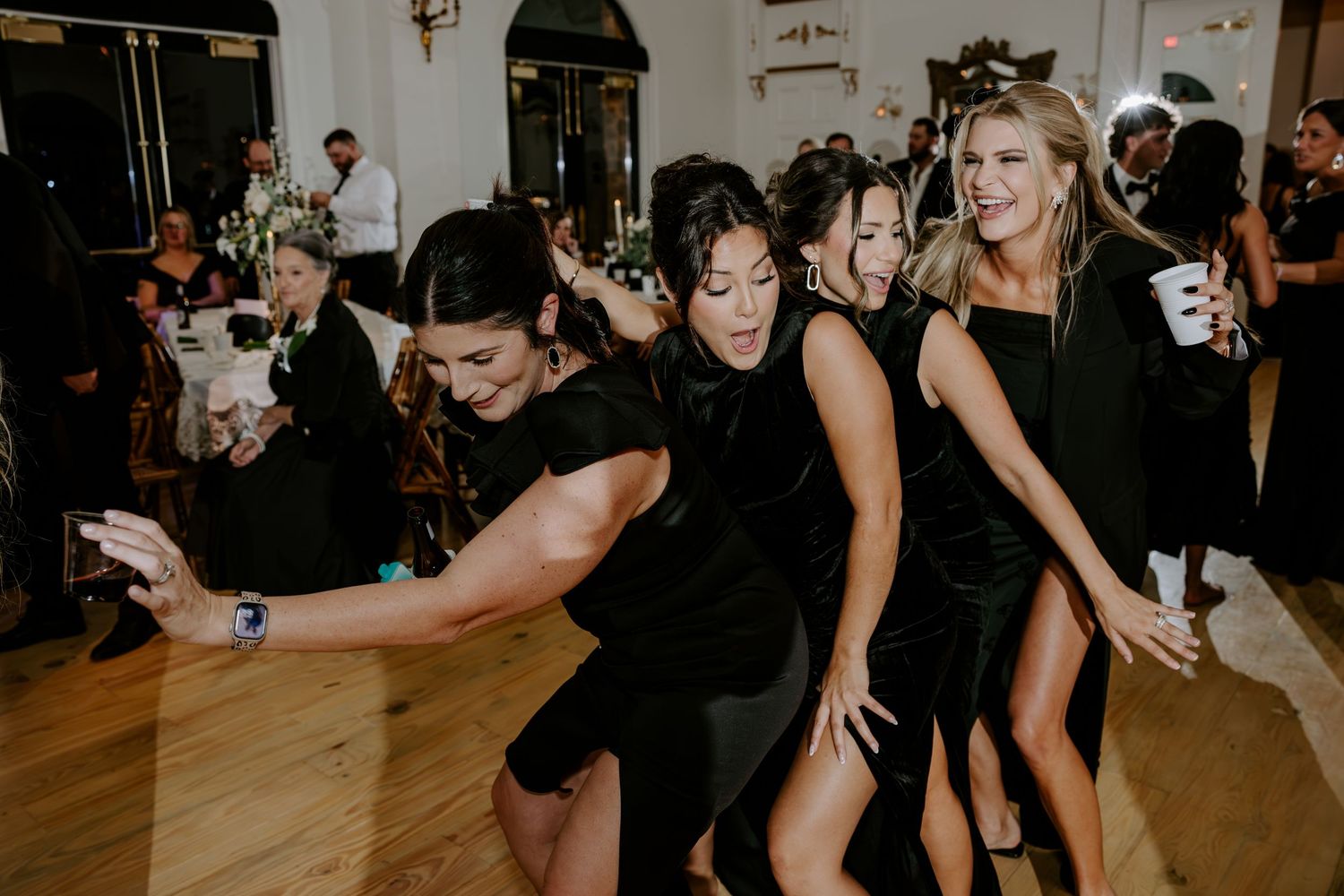Bridesmaids in black dresses laugh and dance together on a wooden dance floor during a lively wedding reception.