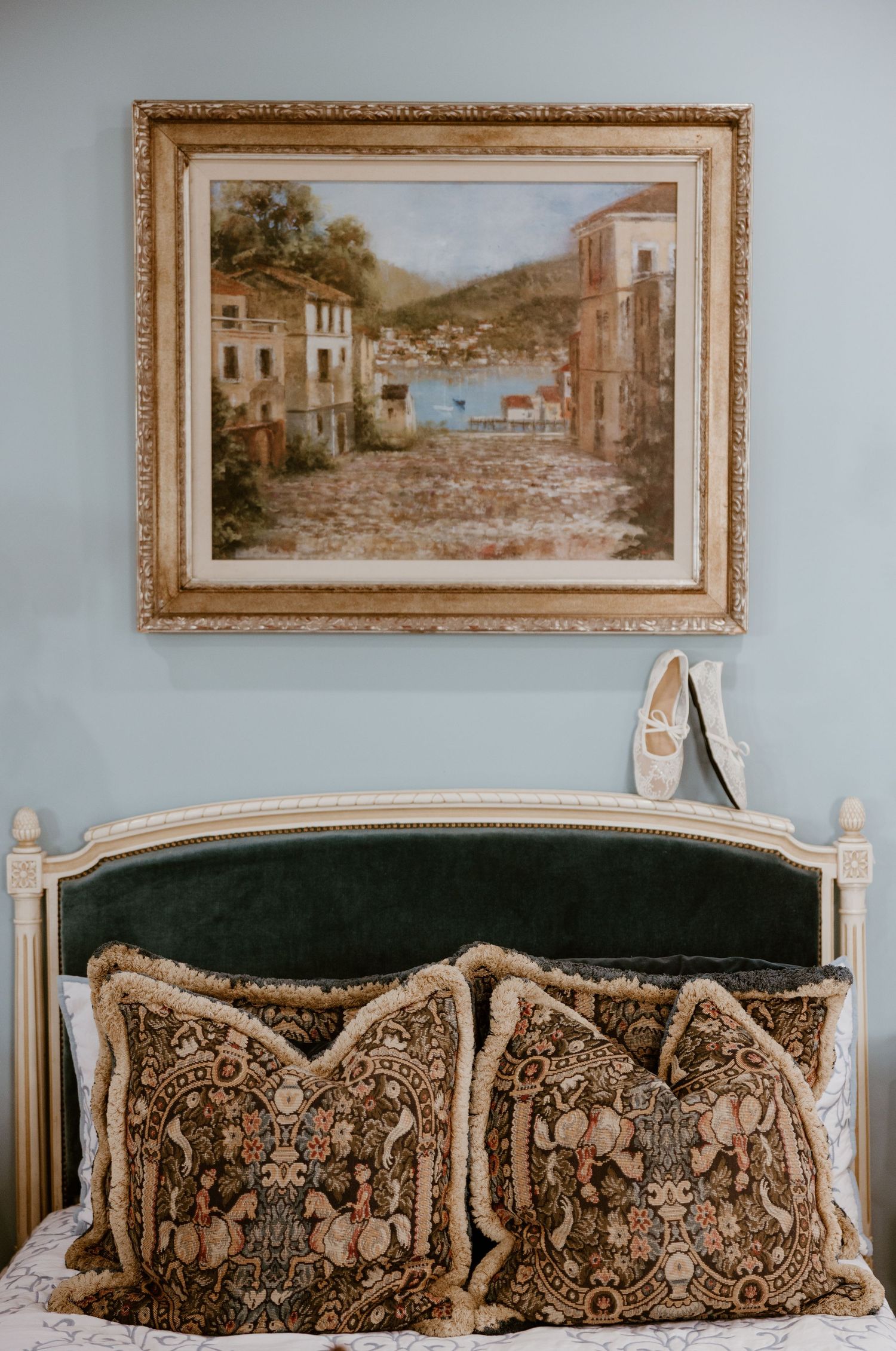 Framed Mediterranean village painting hangs above a green velvet headboard with ornate tapestry pillows and a pair of white flats.