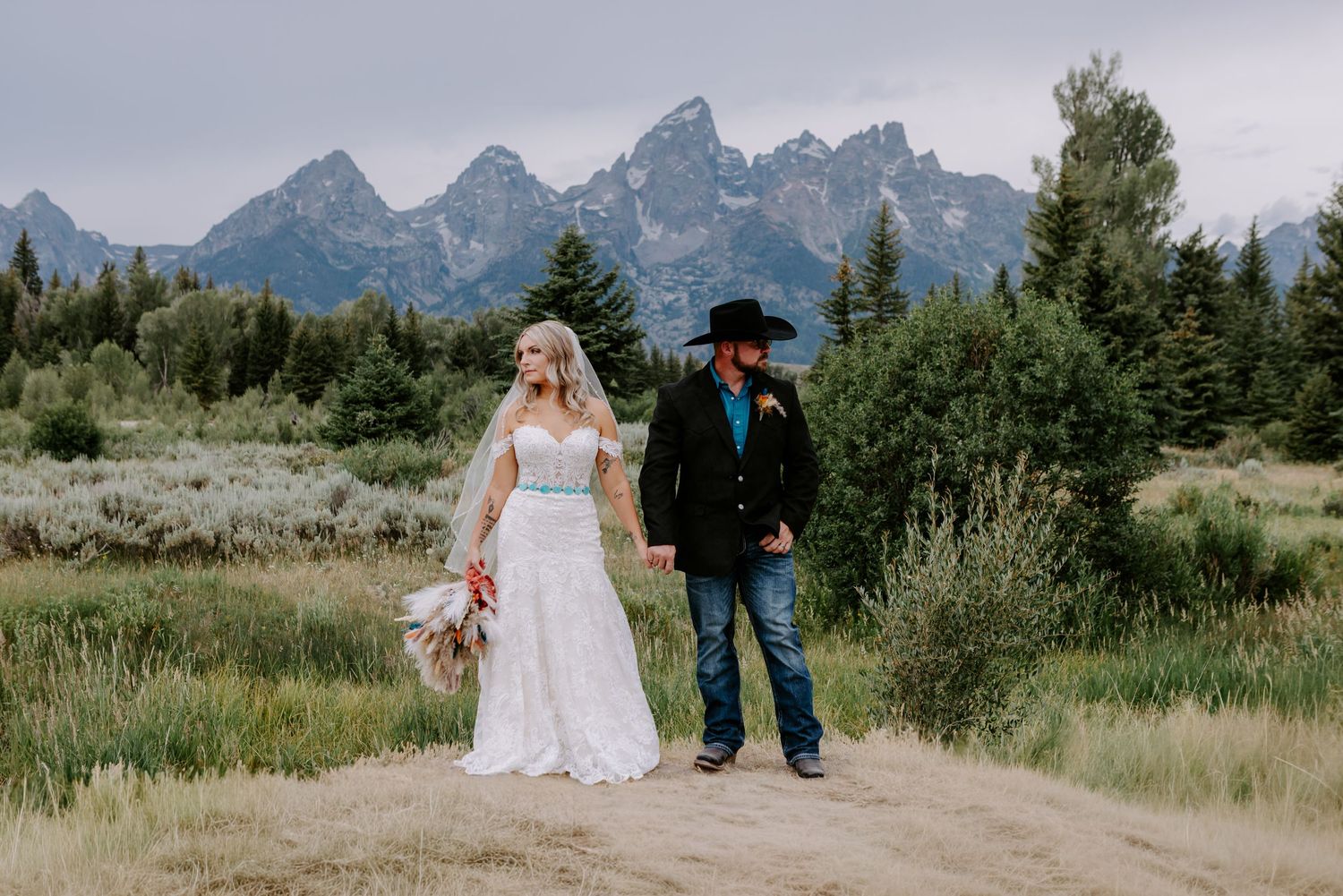 Bride and groom holding hands on a dirt path with Grand Teton mountains and pine trees in the background.
