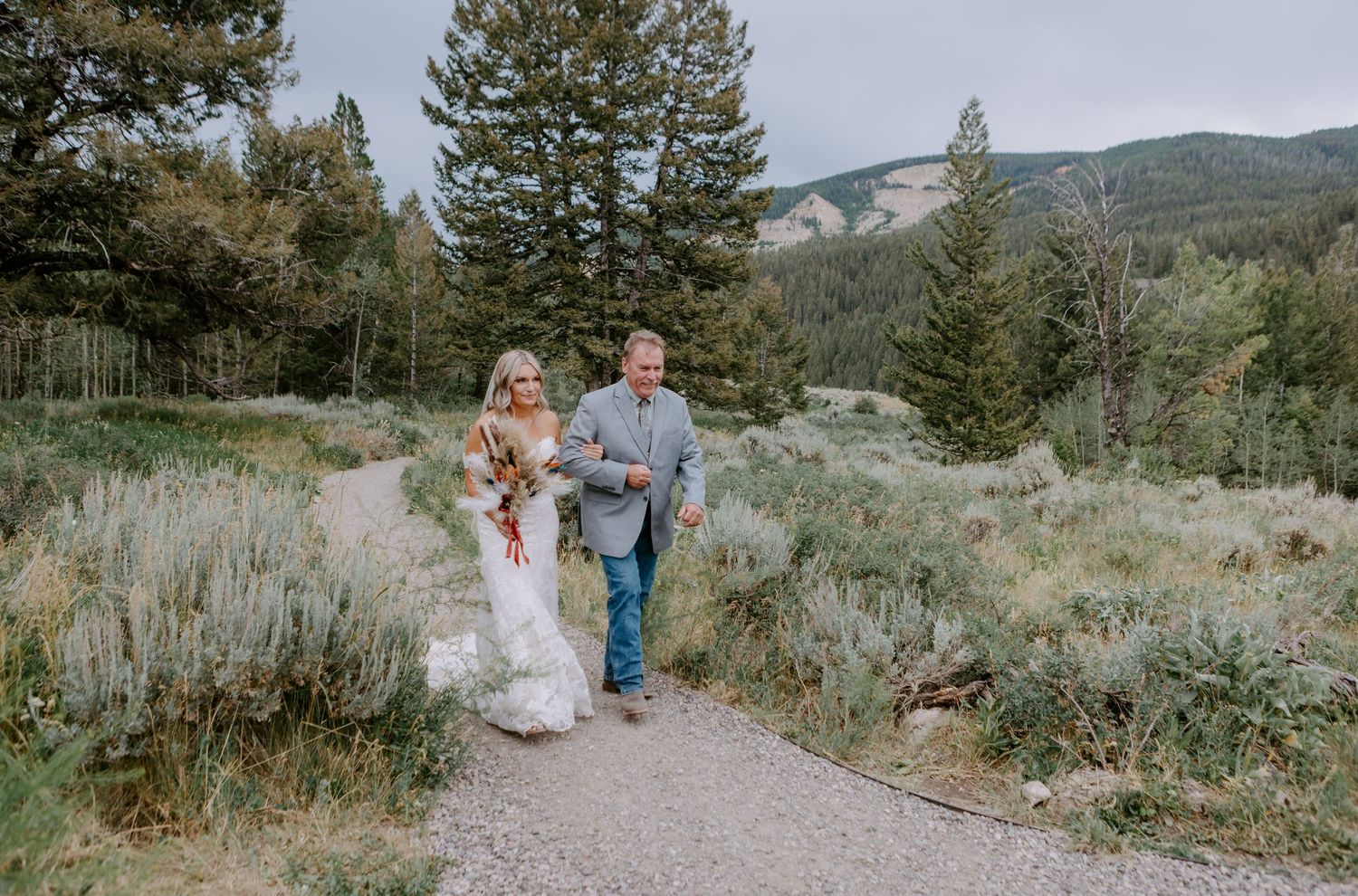 Bride and groom walk hand in hand along a scenic mountain path surrounded by lush greenery and tall pine trees.