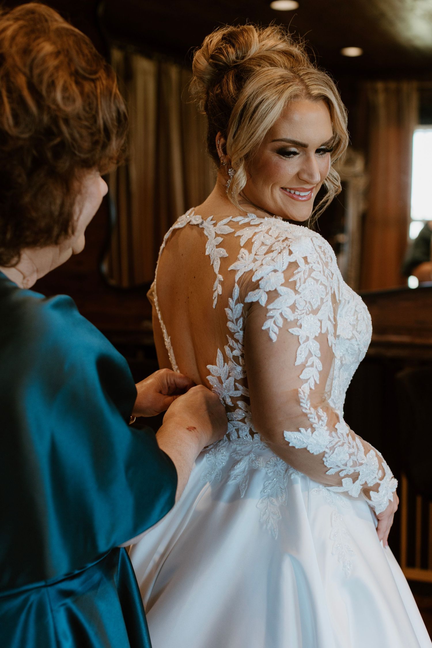 A wedding dress being buttoned up, showing intricate white lace details on the back against a warm wooden interior.