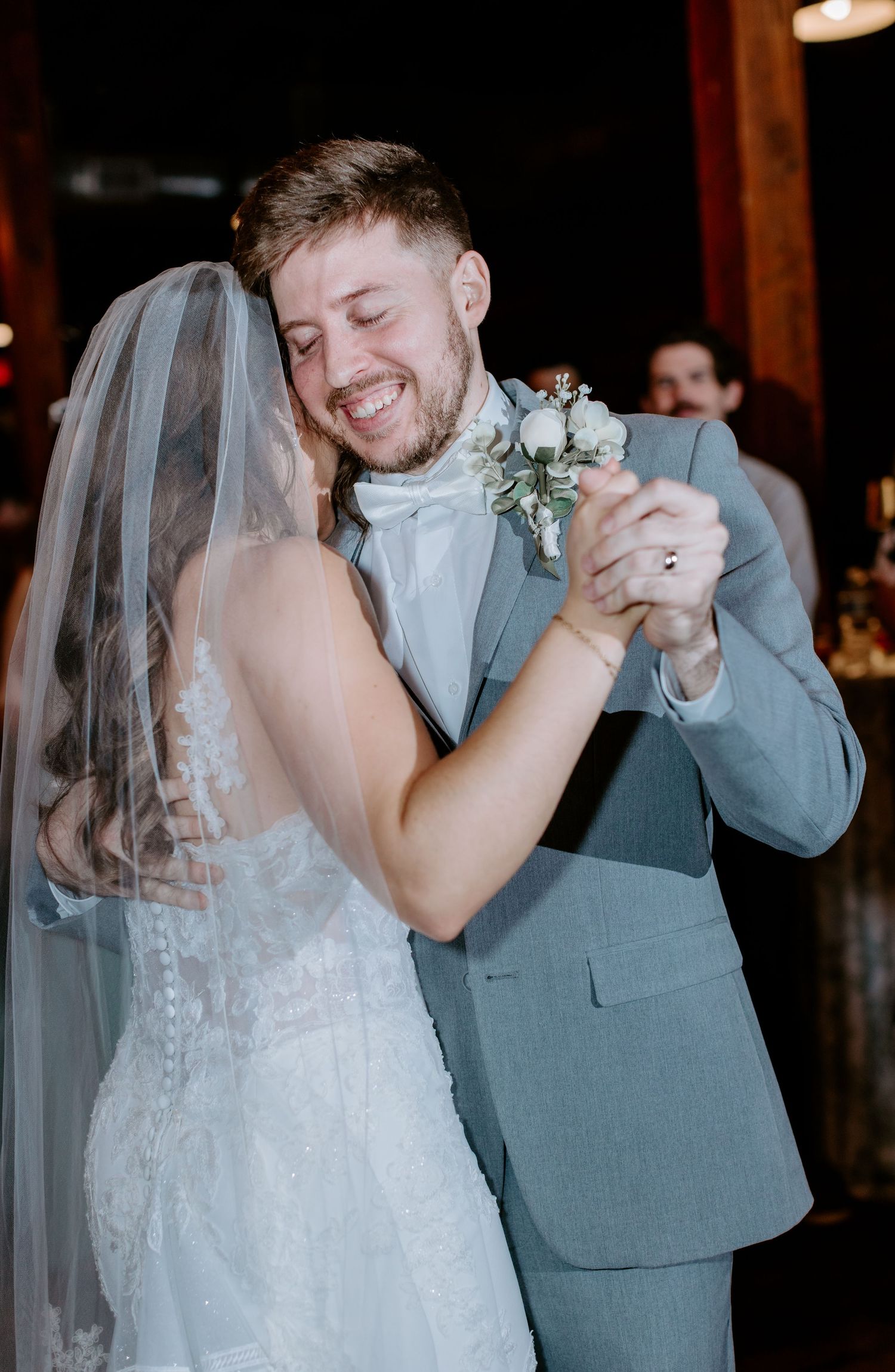 Smiling groom in gray suit shares first dance with bride in white lace wedding dress and veil.