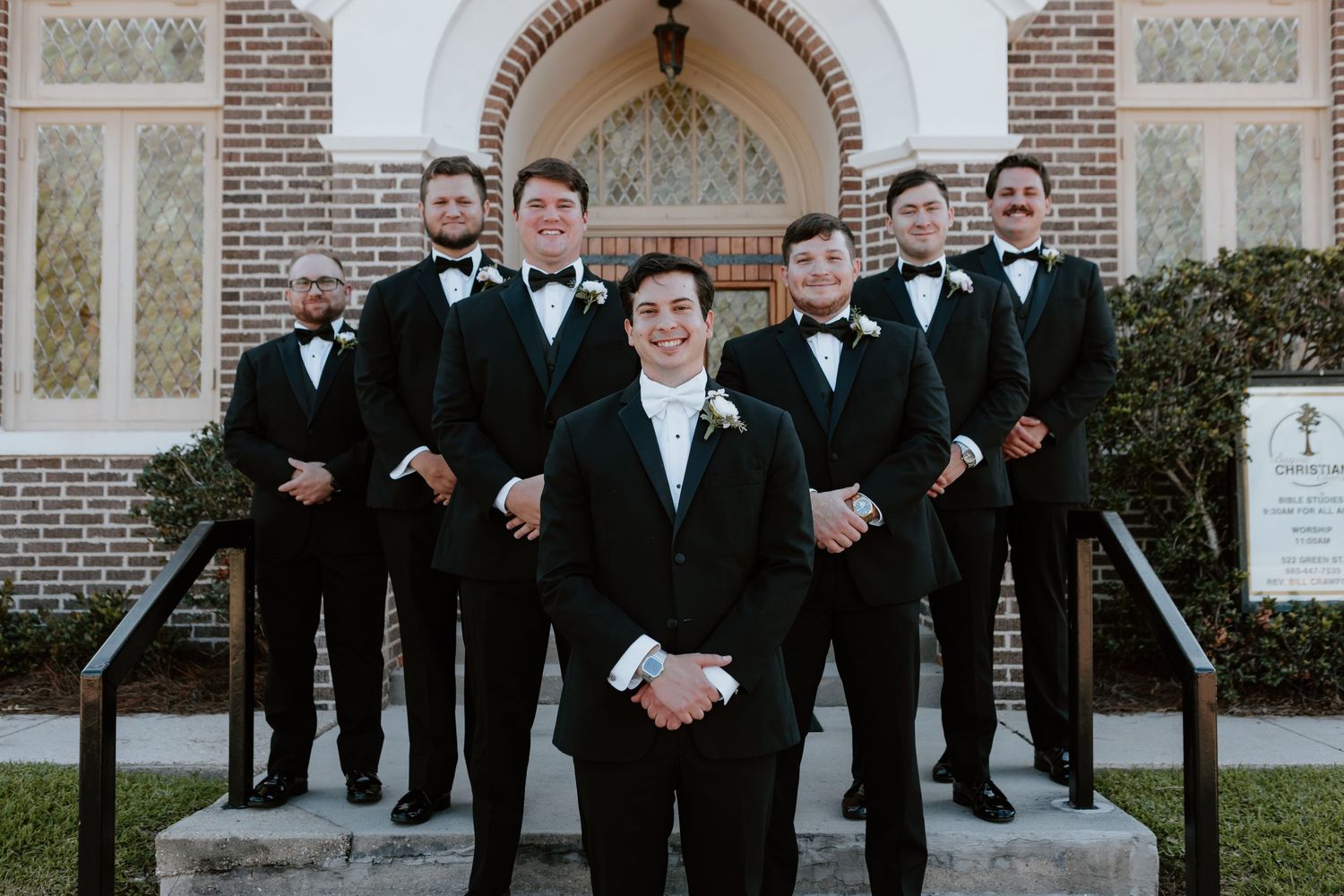 Groom and groomsmen in black tuxedos with bow ties posing on church steps for a classic wedding photo.