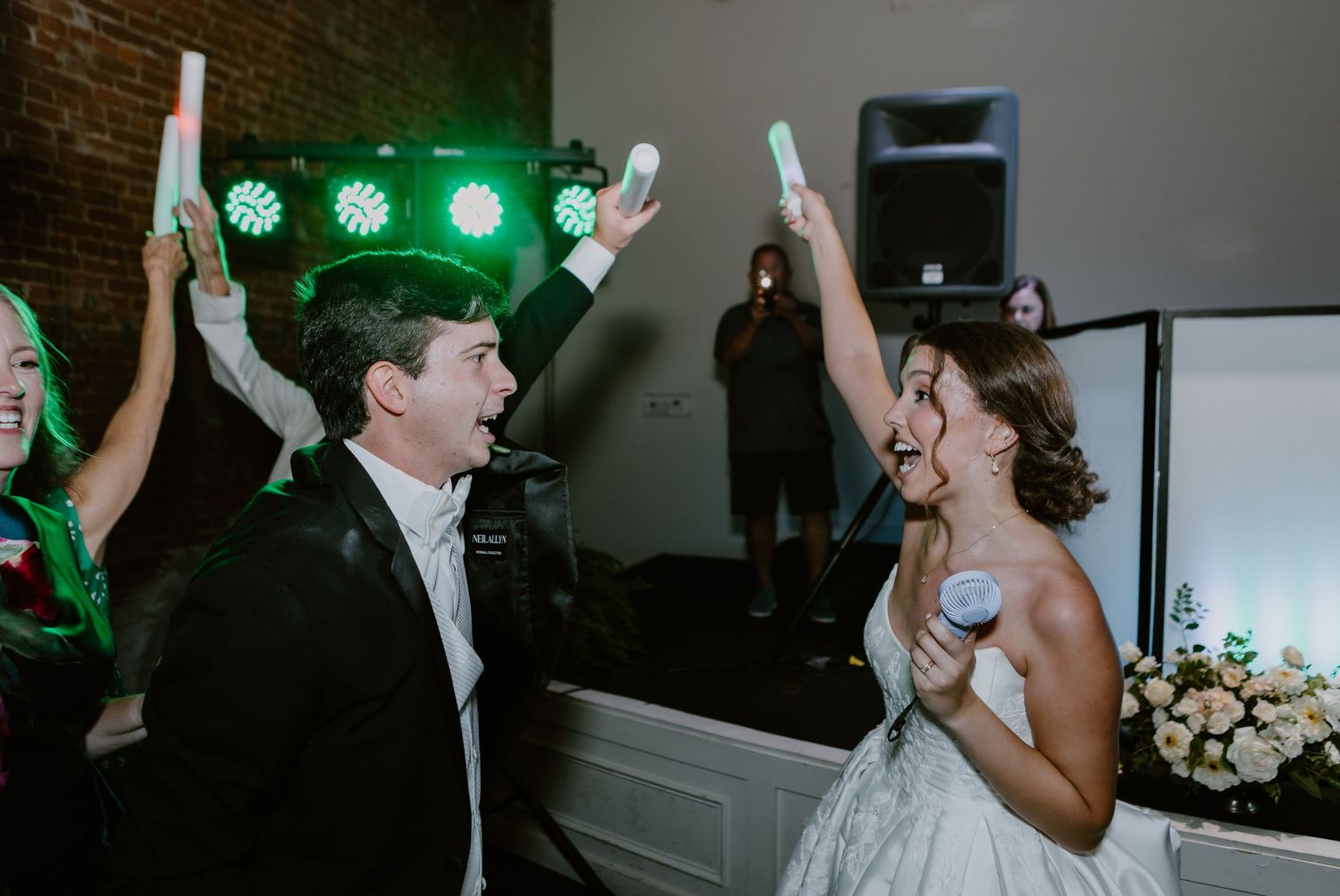 Joyful bride and groom dancing at wedding reception holding glow sticks with DJ lights in background.