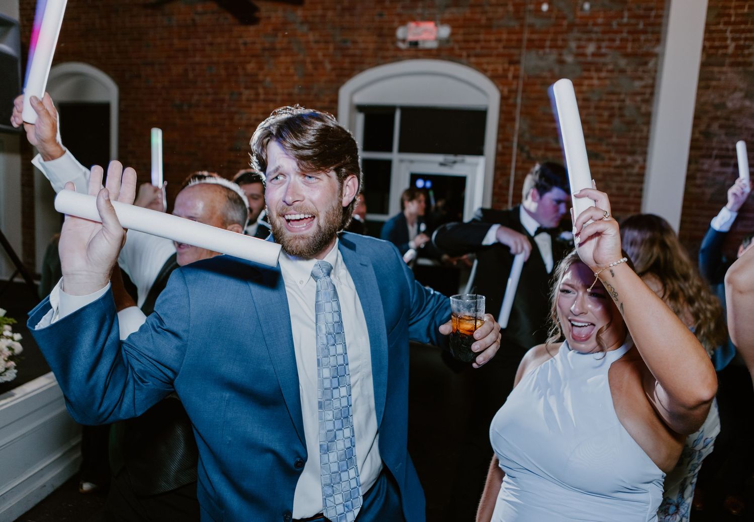 Guests dancing and waving glow sticks at a lively wedding reception in a brick venue.
