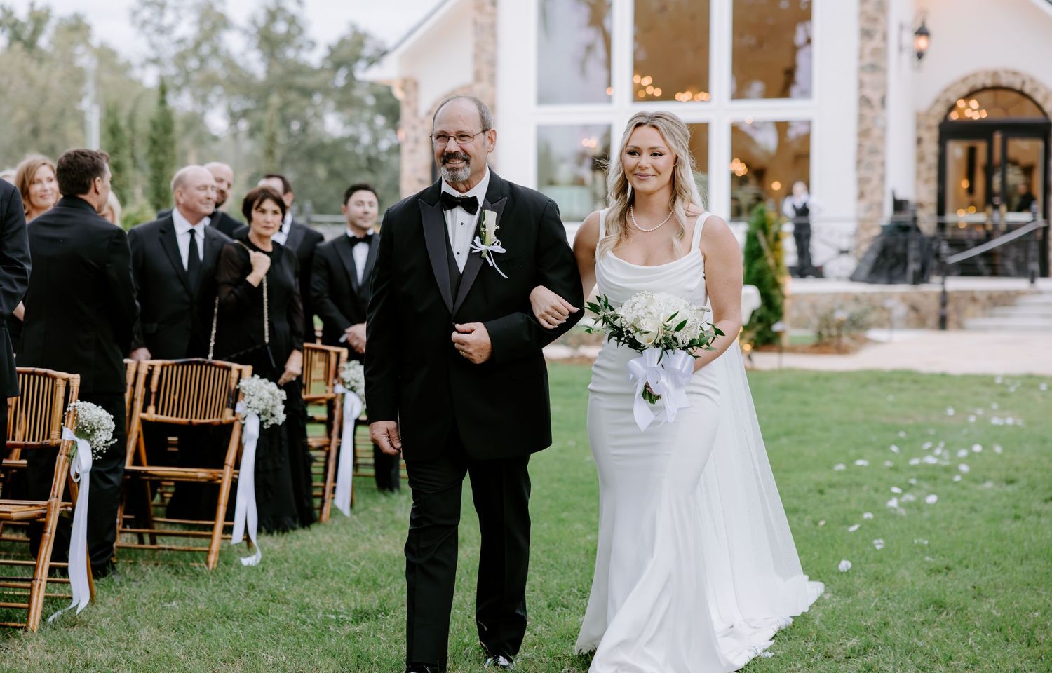 Father in a black tuxedo walks his smiling daughter in a lace wedding gown down the outdoor aisle past seated guests at a garden ceremony.
