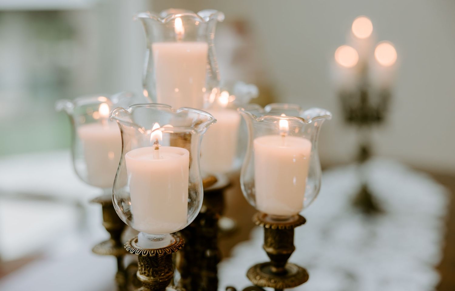 White pillar candles glowing inside clear glass hurricane holders on antique brass candlesticks, creating a warm, elegant table centerpiece.