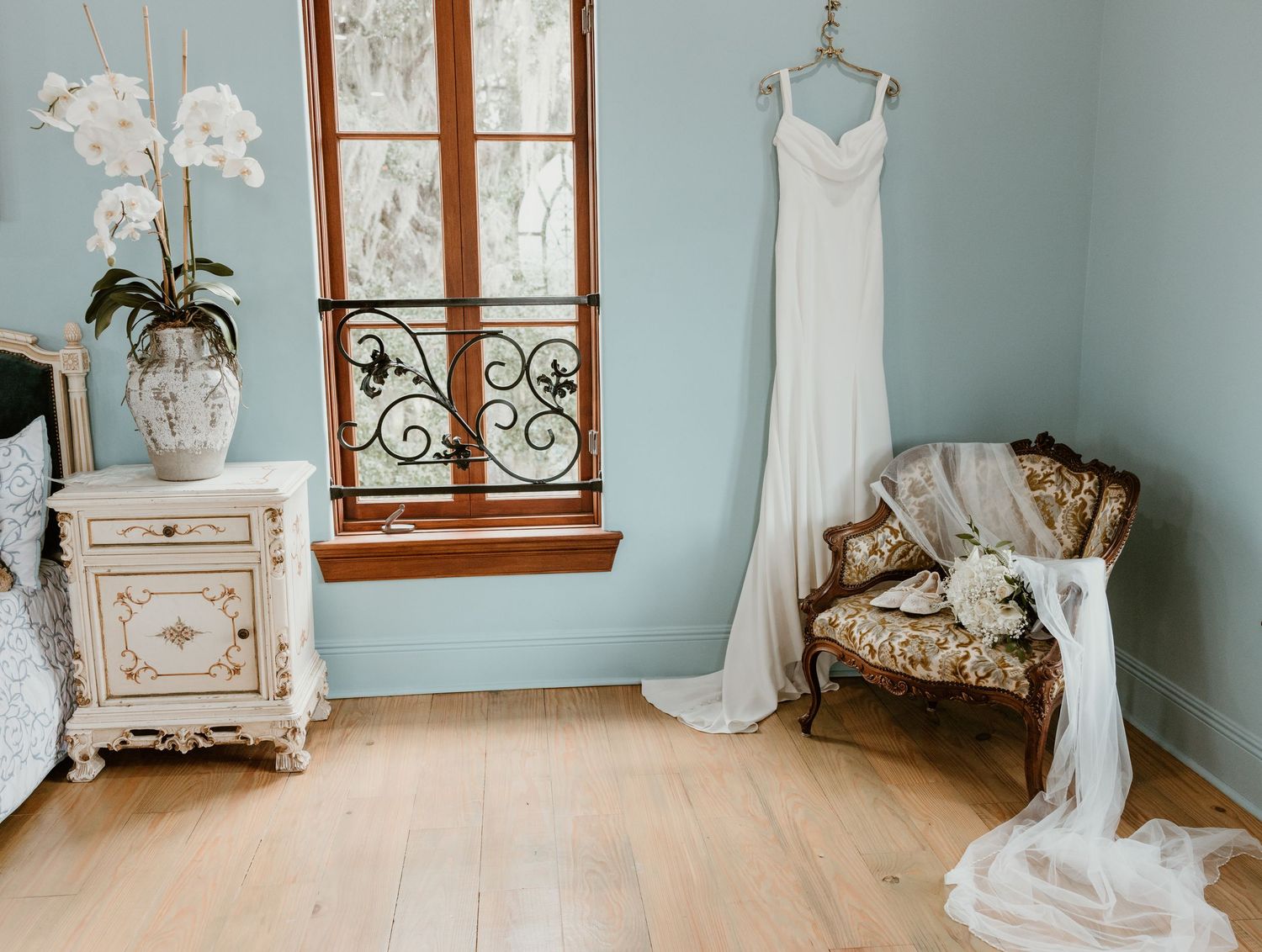 White wedding dress hanging by a window above a vintage armchair with a bridal bouquet in a soft blue room with an ornate white nightstand.