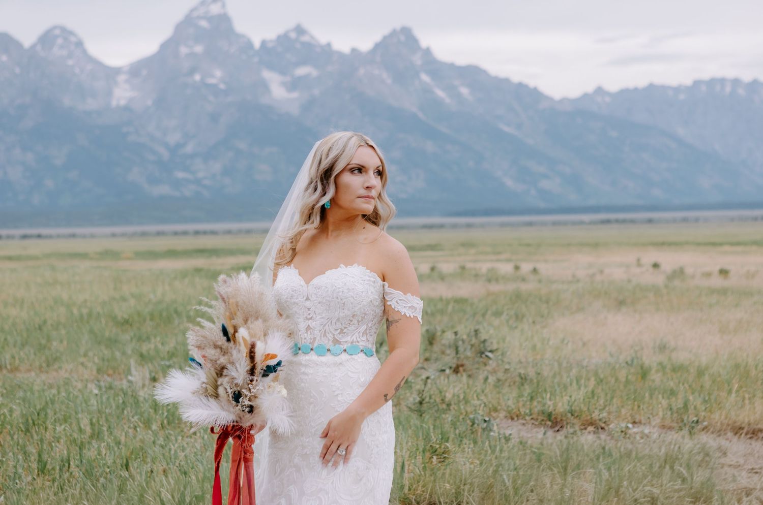 Bride in off-shoulder lace gown holding bouquet with Grand Teton mountains backdrop in Wyoming.