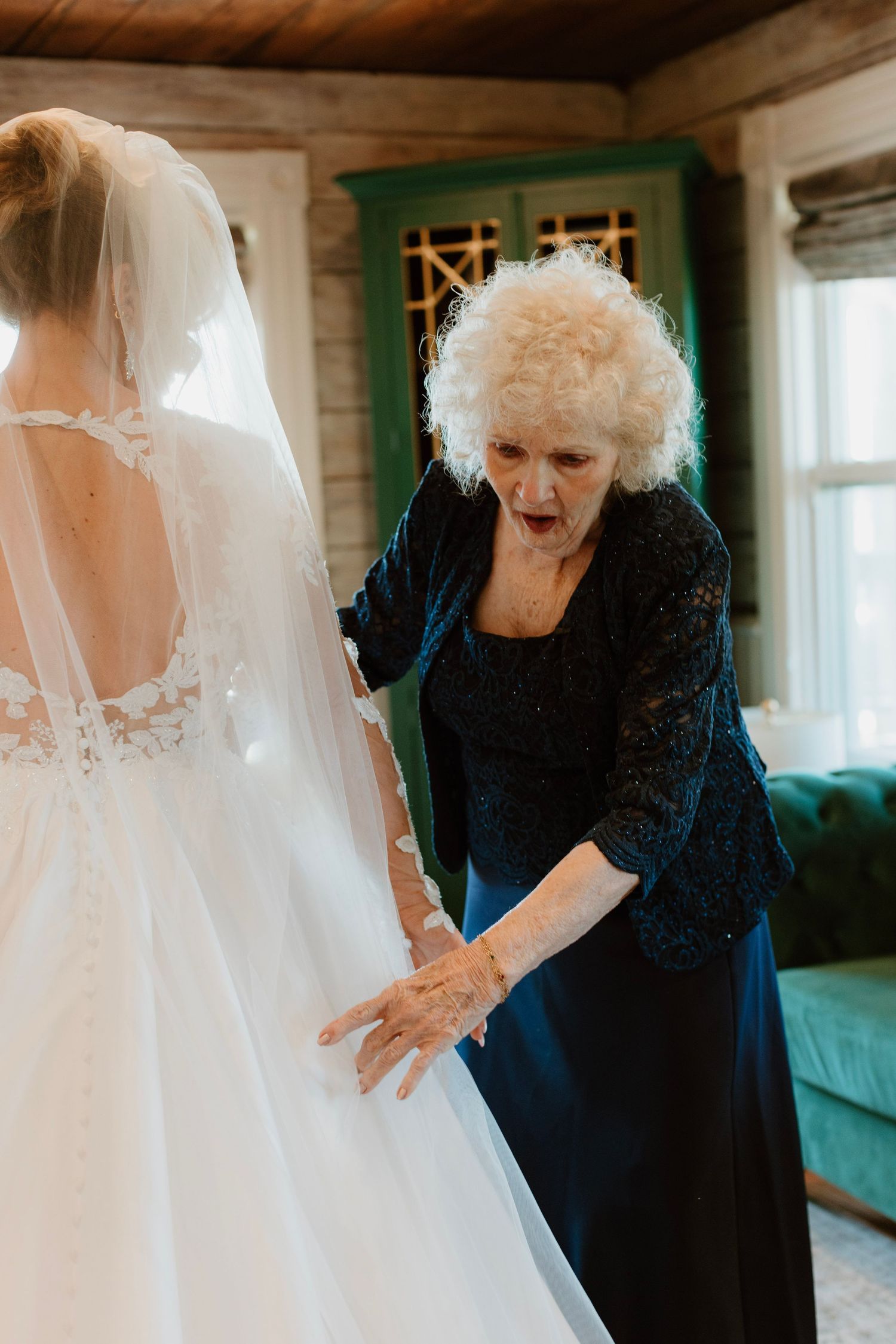 Elderly woman in navy dress reacts with awe to bride in white lace wedding gown and veil indoors.