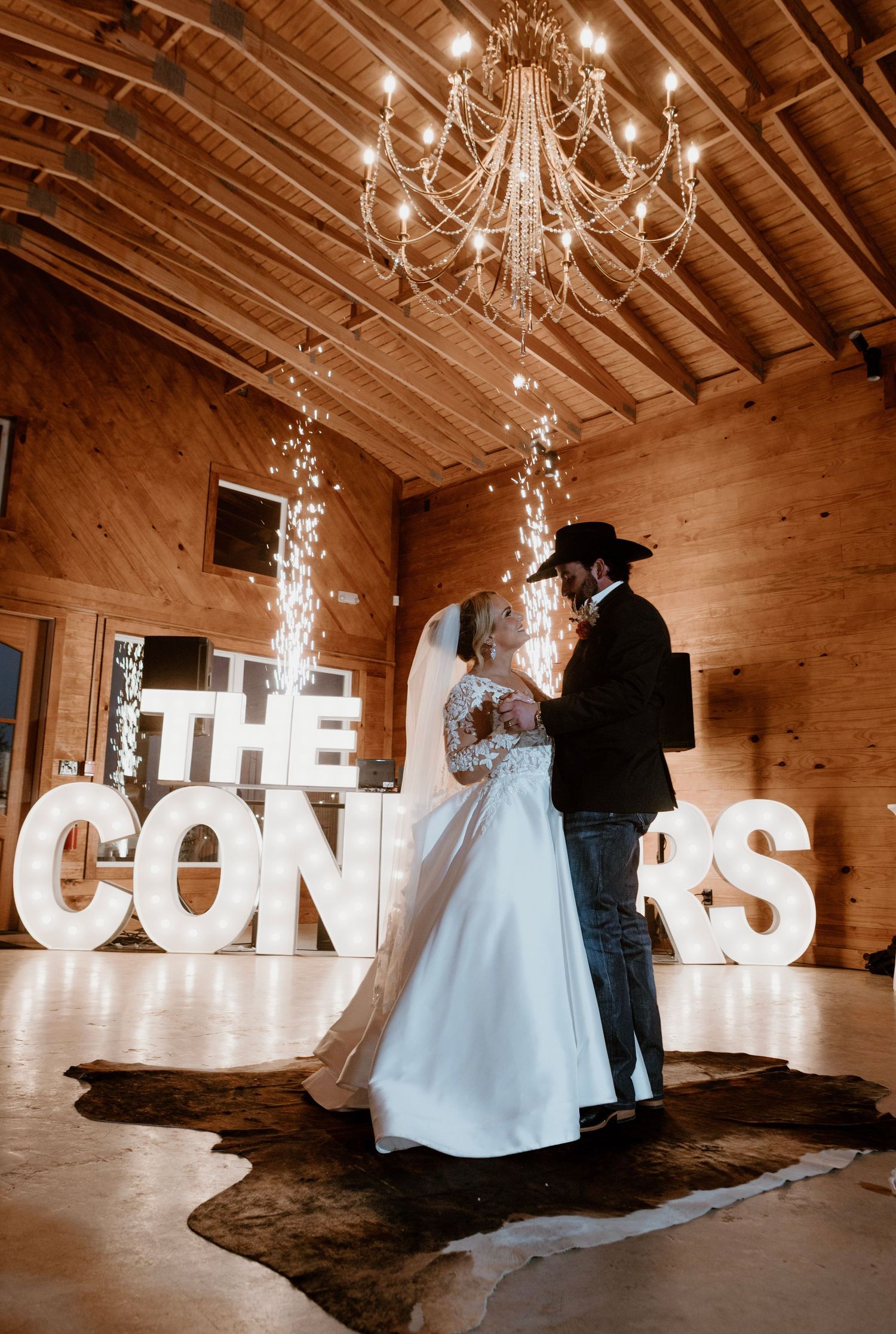 Newlyweds share first dance under chandelier beside illuminated THE CONNERS marquee letters.