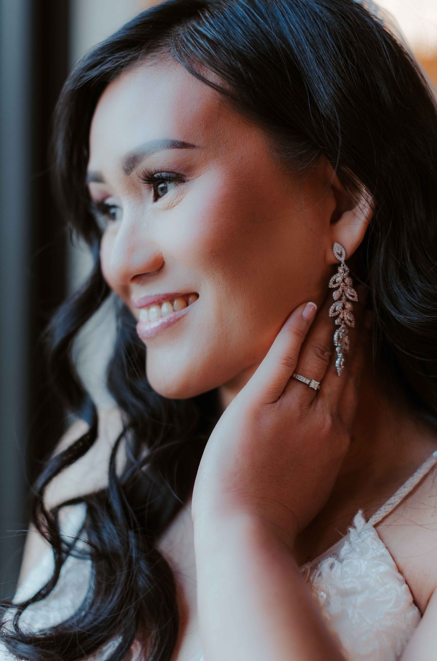 A bride with long dark hair wearing crystal drop earrings and a white wedding dress smiles in soft light.
