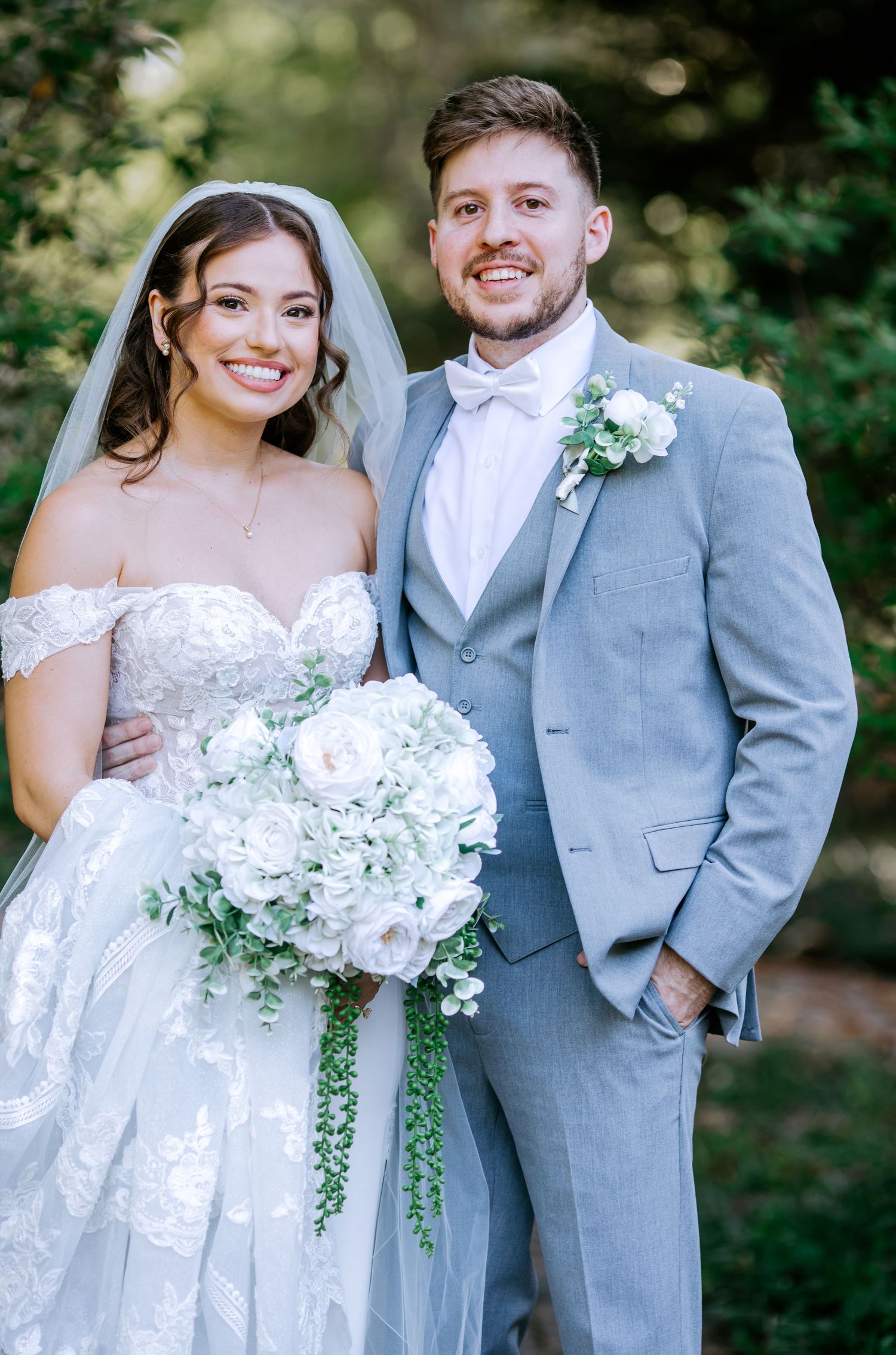 Happy bride and groom pose together outdoors, she in lace off-shoulder gown with white bouquet, he in gray suit.