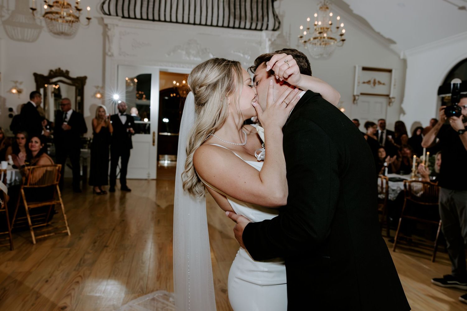 Bride in a white gown and long veil kisses her groom in a black suit on the dance floor as wedding guests watch in a elegant venue.