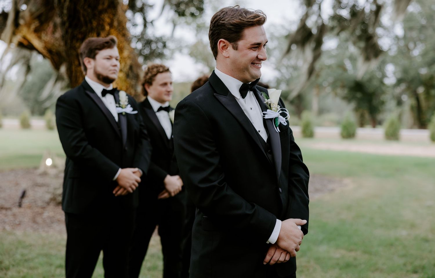 Smiling groom in a black tuxedo with bow tie stands outdoors at a wedding ceremony while groomsmen in matching suits look on.