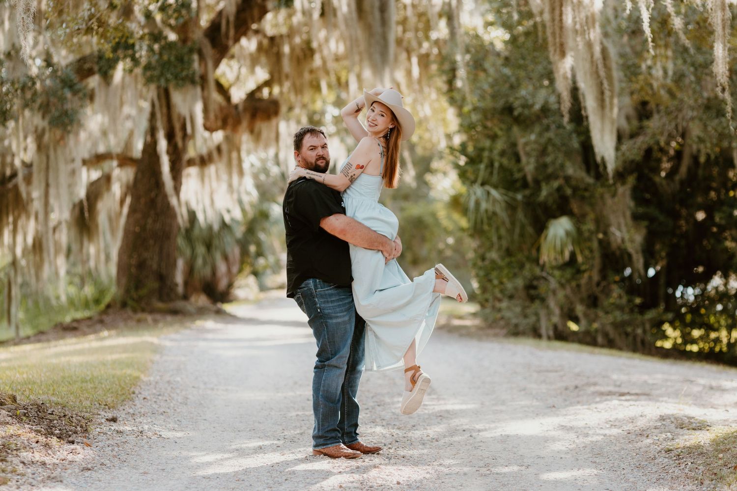 A couple shares a romantic moment on a tree-lined path with Spanish moss hanging overhead during golden hour.
