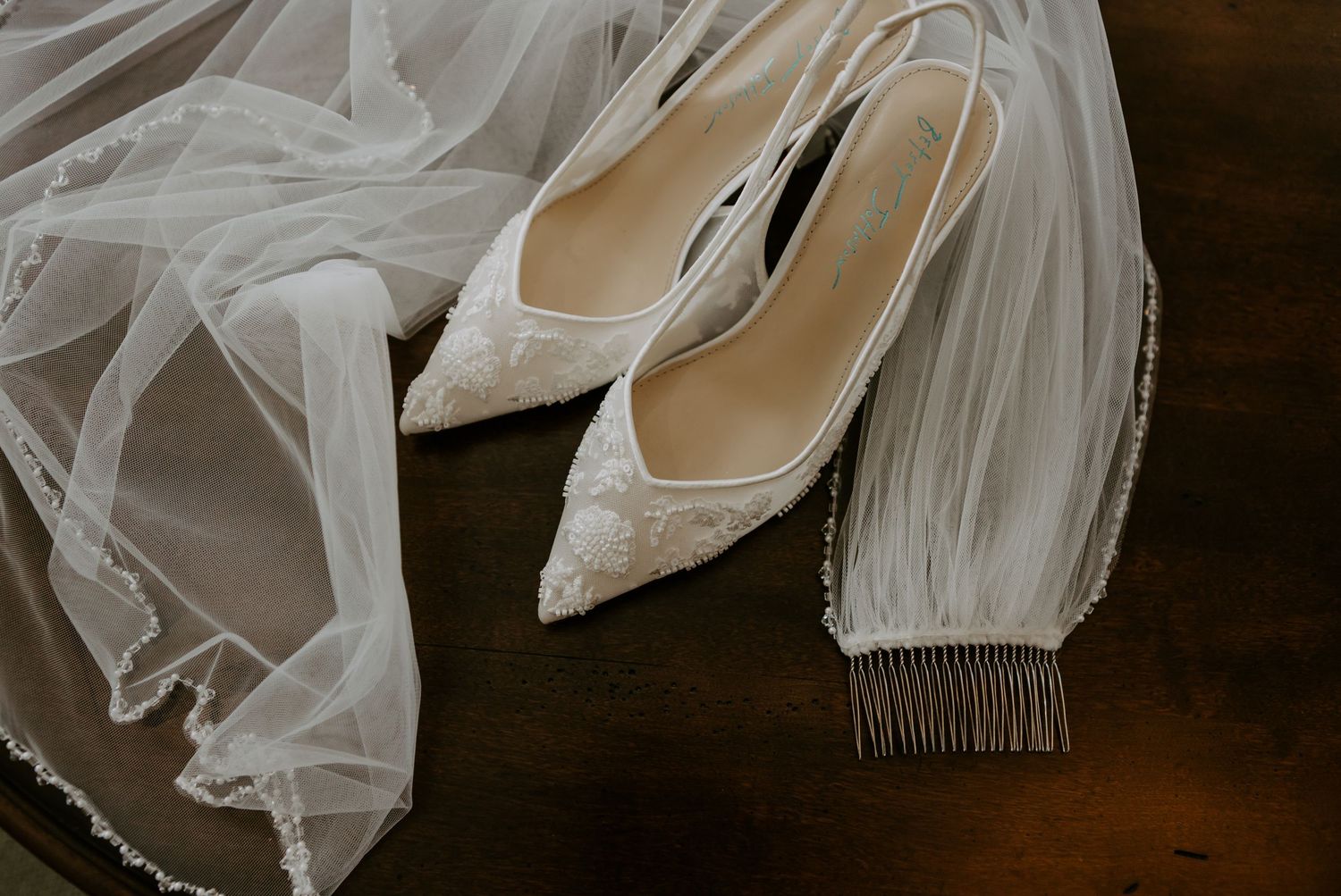 Elegant white lace bridal heels with fringe detail resting beside a sheer wedding veil on a wooden surface.