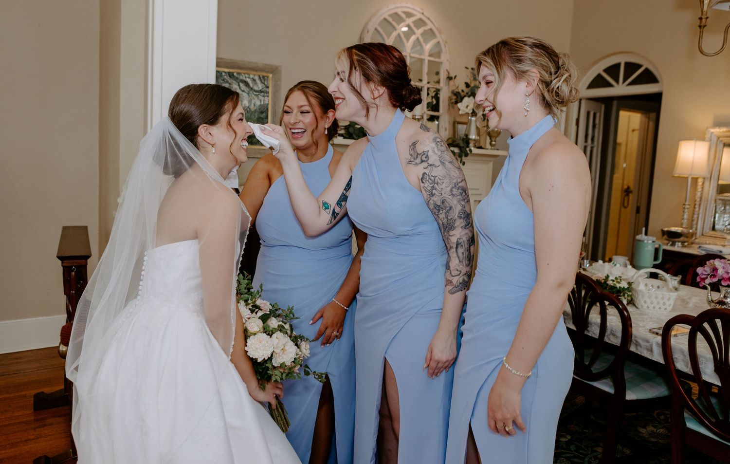 Bride in white gown and veil greeted by bridesmaids in blue dresses holding bouquet.
