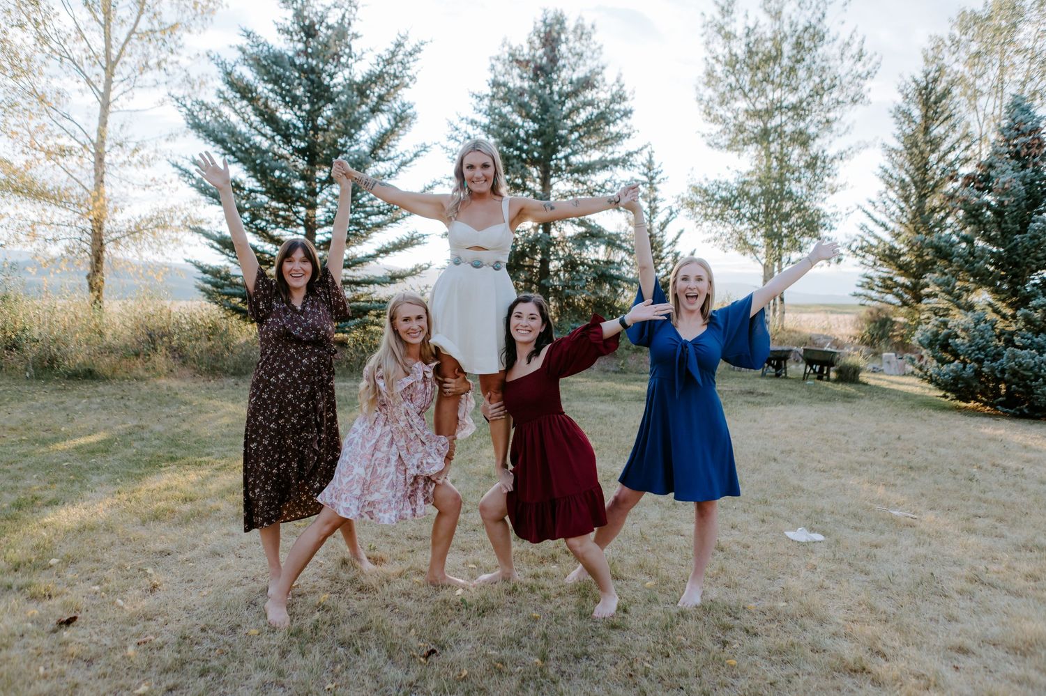 Bride in white dress lifted by four bridesmaids in colorful dresses outdoors with pine trees in background.