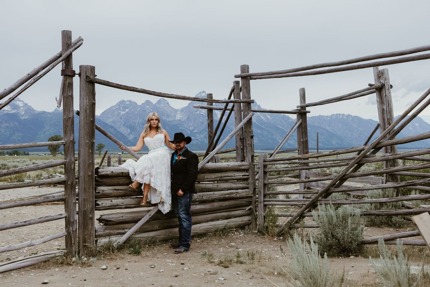 Bride in white dress sits on rustic wooden fence with groom in cowboy hat, Grand Teton mountains backdrop.