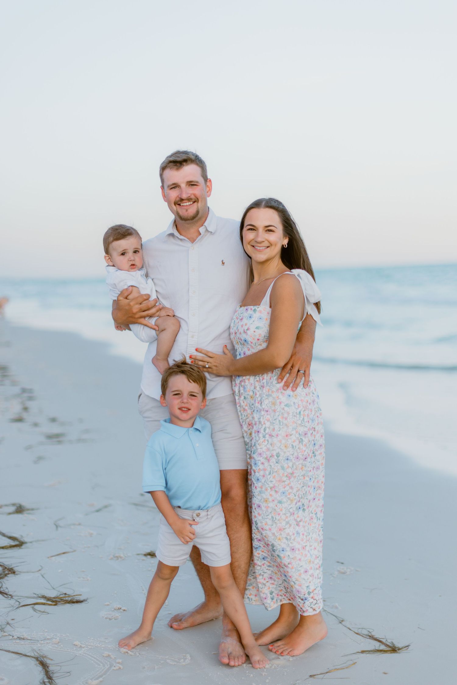 Family in coordinated beach outfits poses together on white sand beach at sunset with ocean waves in background.