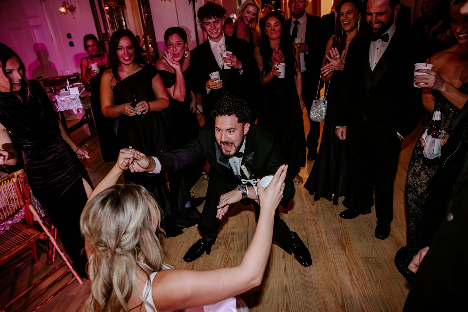Wedding guests in formal attire cheer and clap as a groom in a tuxedo dances with a blonde bride on a wooden dance floor at the reception.