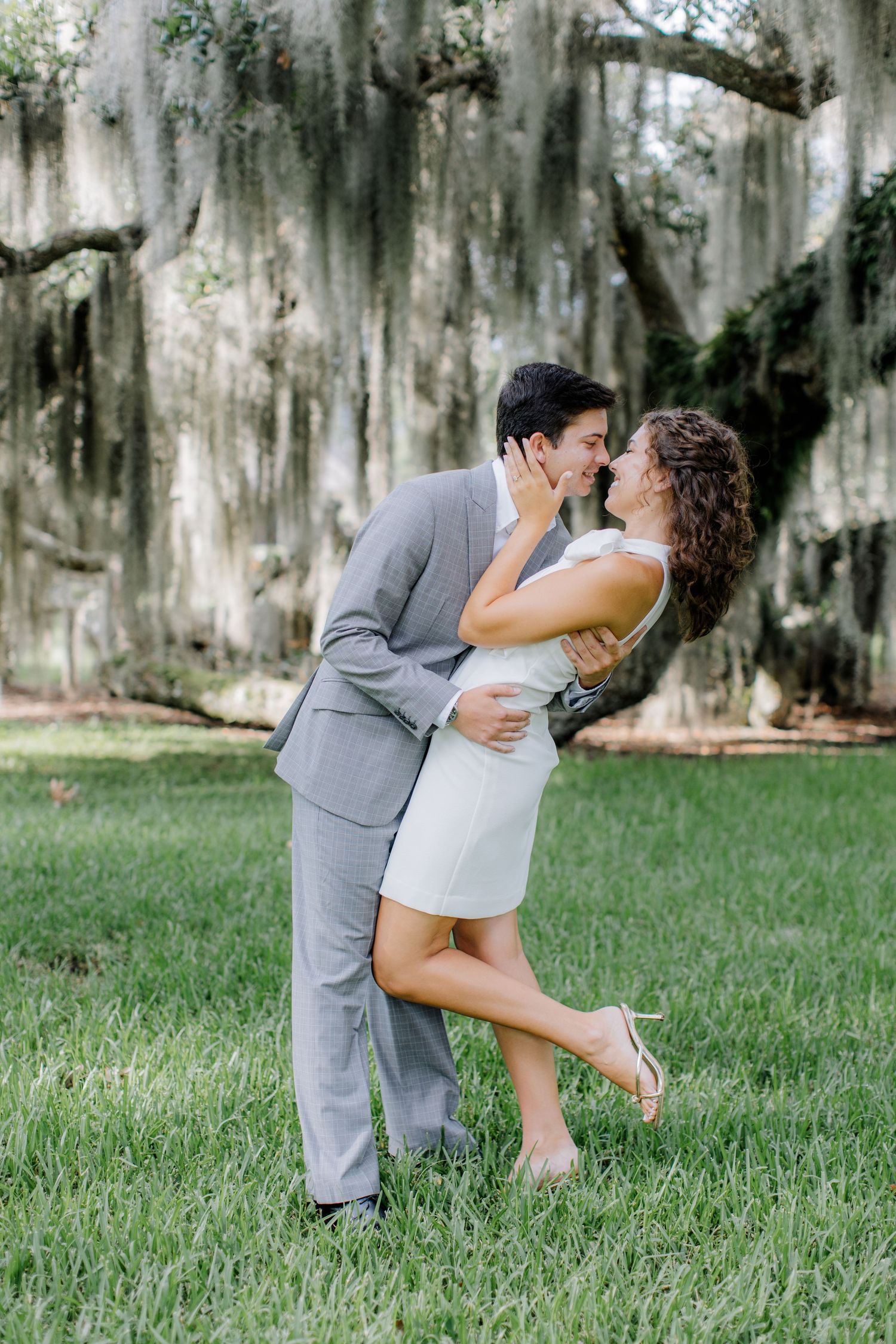Romantic couple in grey suit and white dress dance and embrace on grass beneath Spanish moss draped trees.