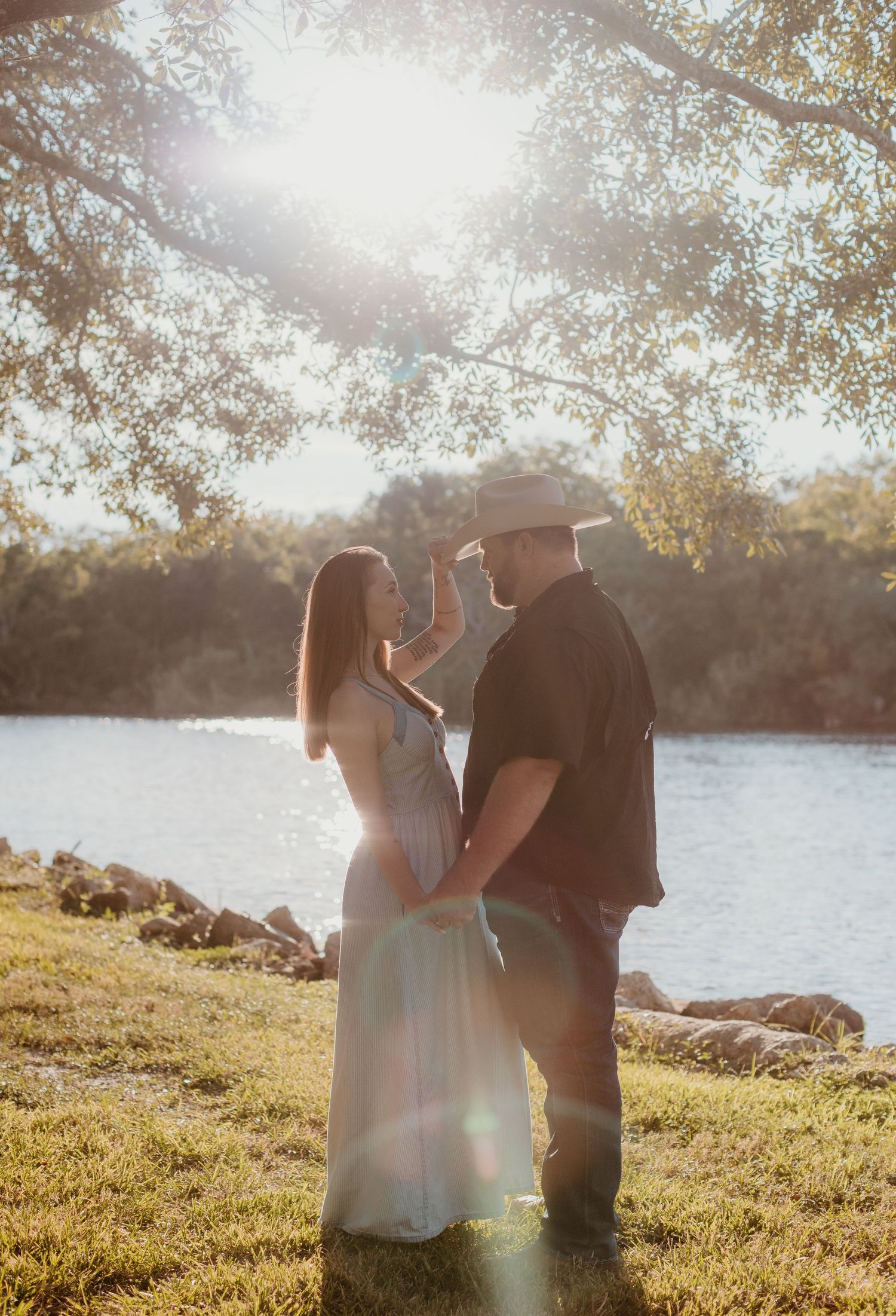 A couple shares a romantic moment by a sunlit lake on a grassy shore with lens flare effects in the background.