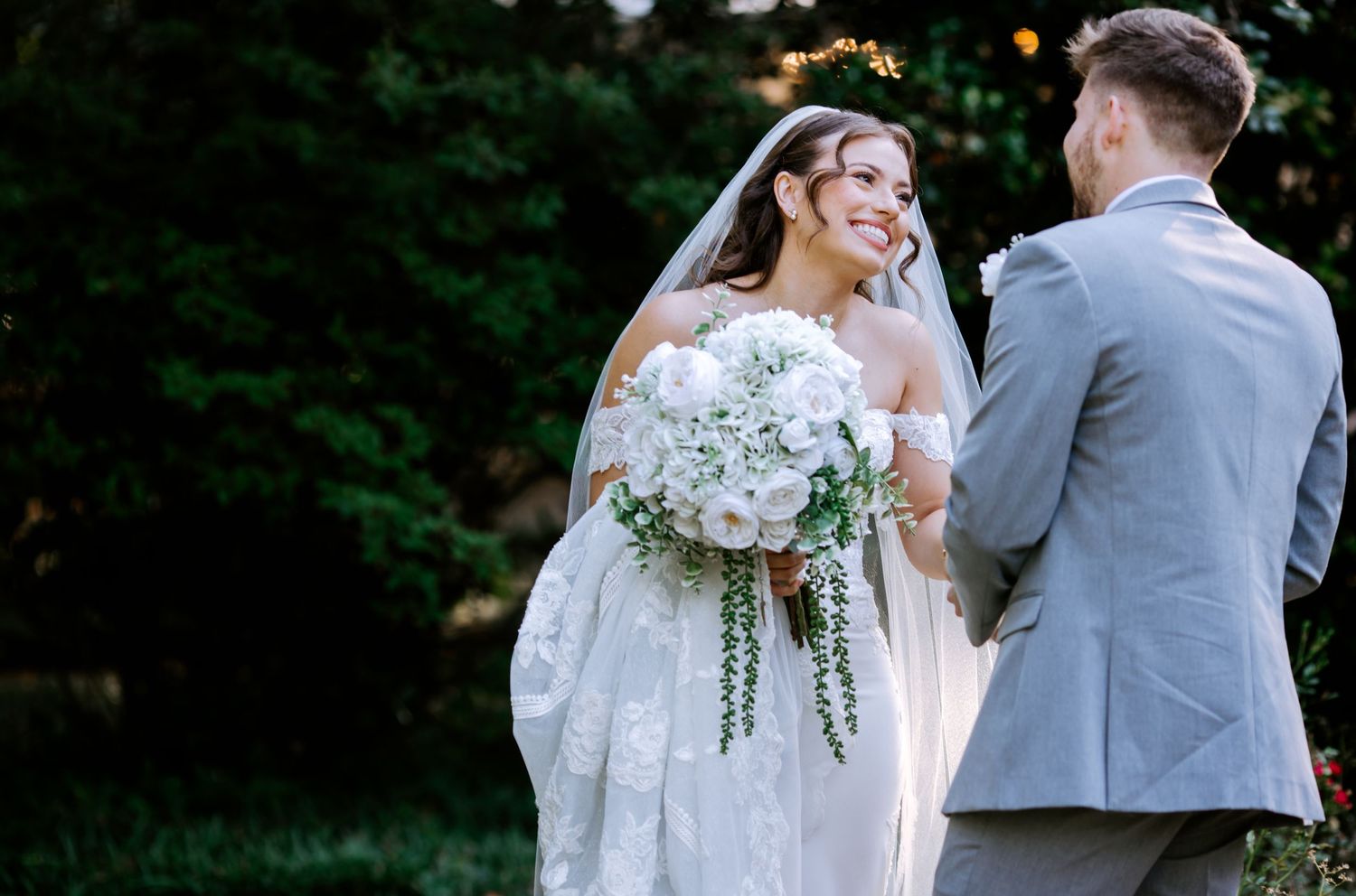 Smiling bride in white lace gown holding white floral bouquet with groom in gray suit outdoors.
