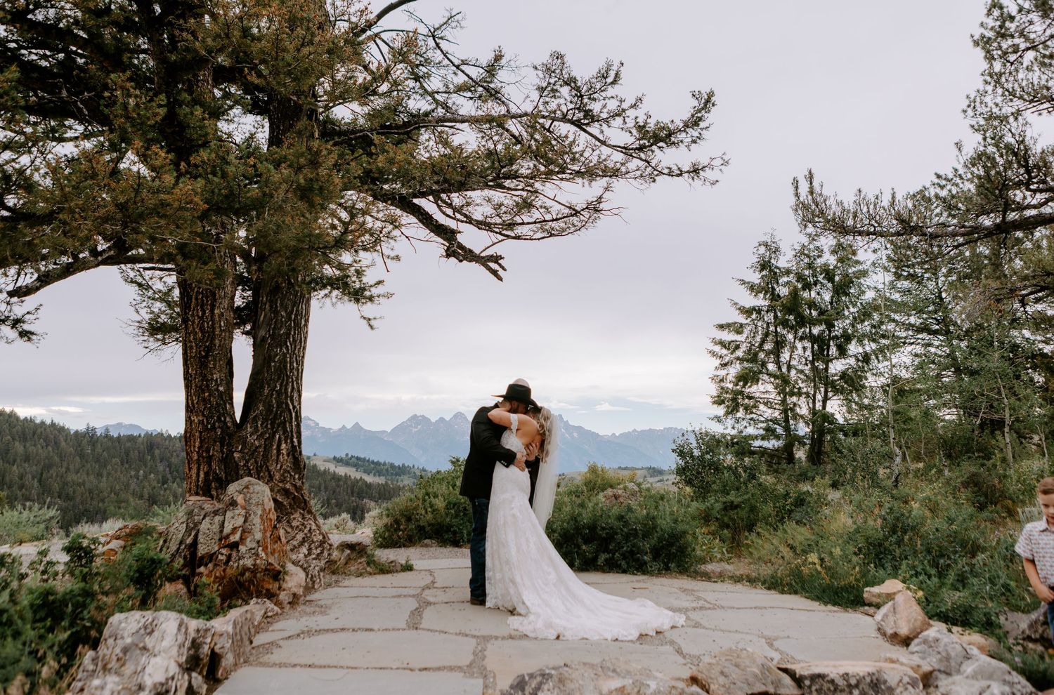 Bride and groom kissing on stone terrace with mountain views and pine trees at outdoor wedding.