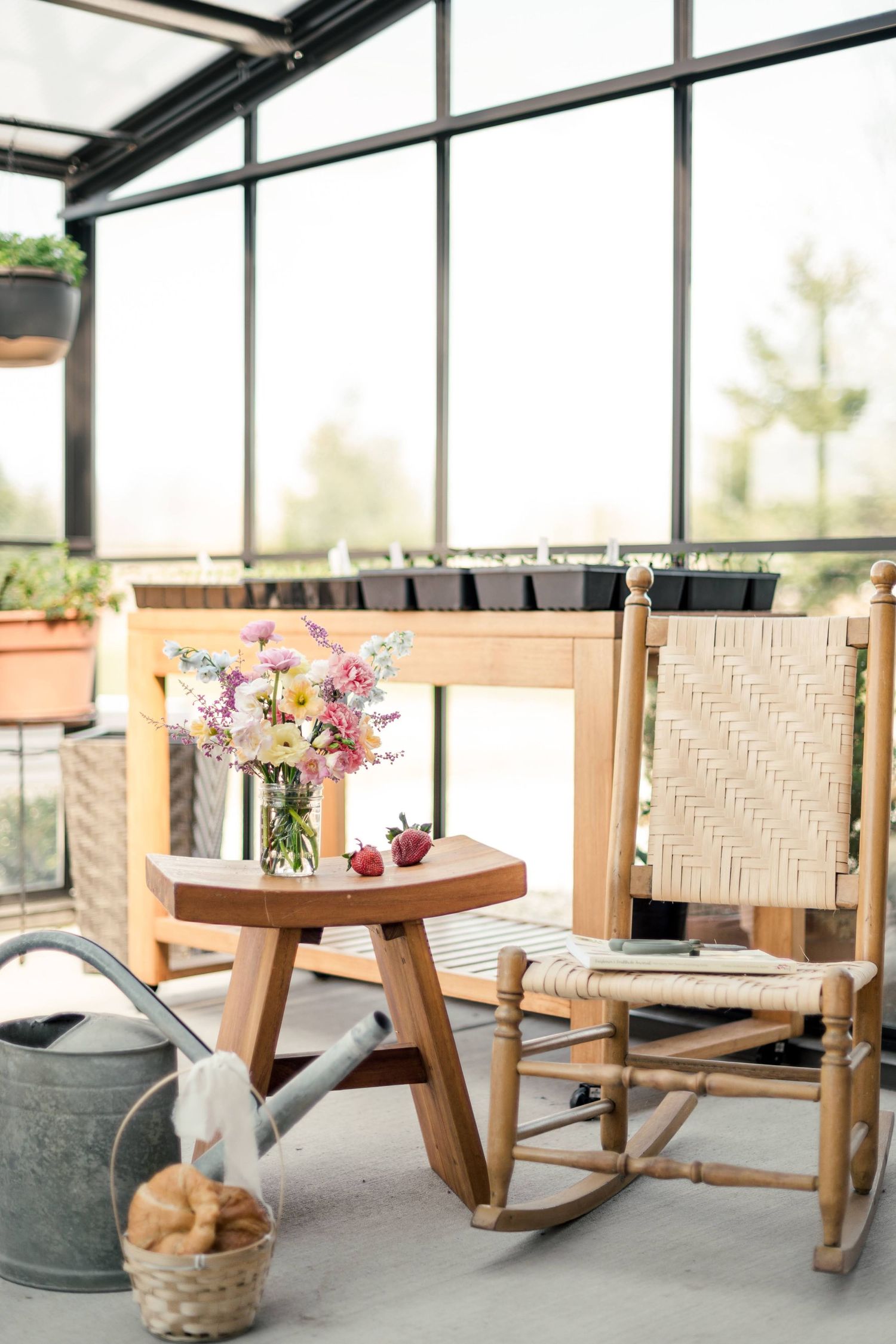 Cozy sunroom corner with woven rocking chair, wooden side table holding fresh flowers, and vintage watering can by window.